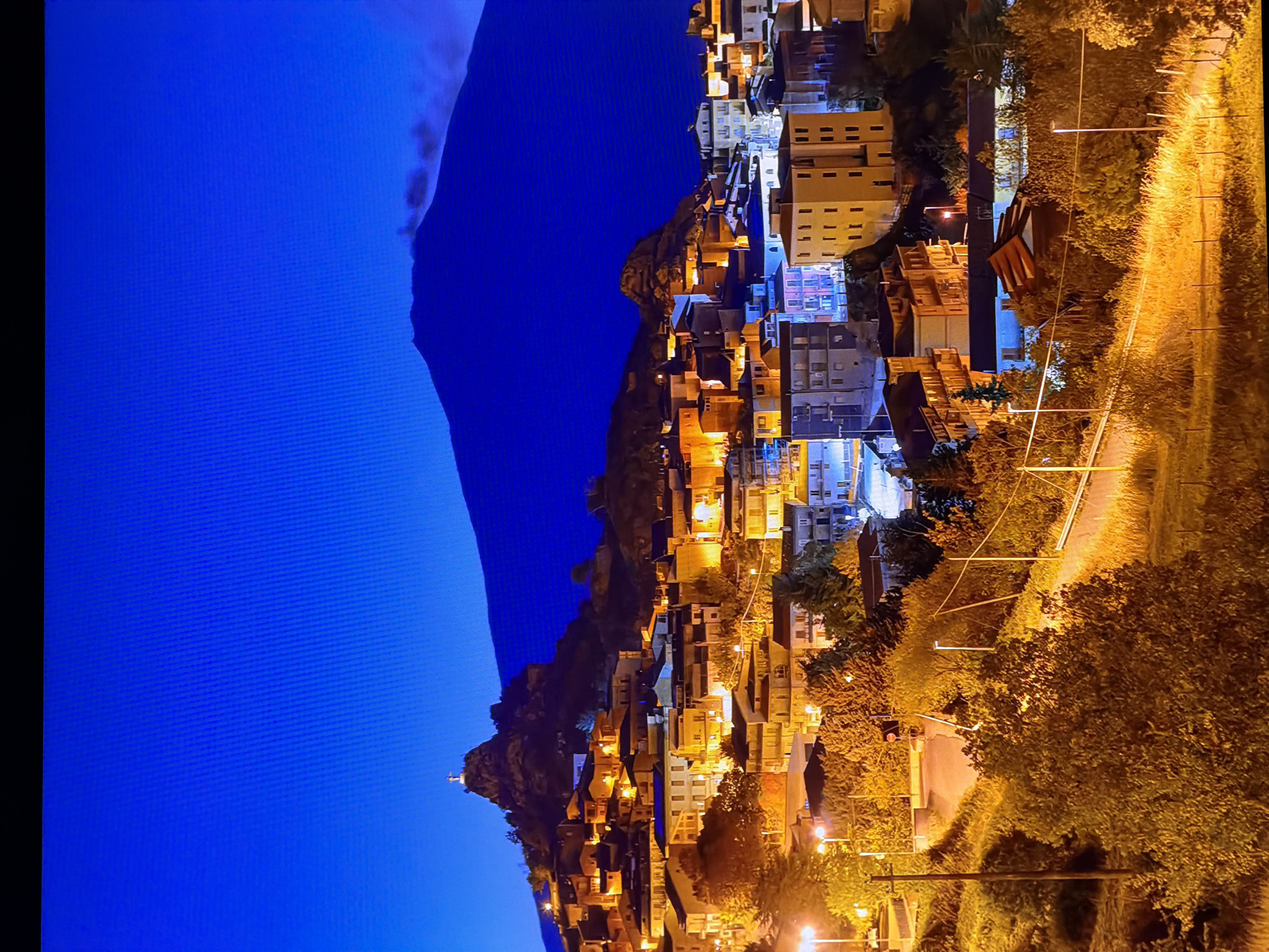 The image depicts a nighttime scene of a town nestled at the base of a large mountain, likely a volcano, under a deep blue sky. The town is illuminated by numerous lights, highlighting the architecture of the buildings, which appear to be made of stone and plaster. The houses are densely packed, with some situated on steep hillsides, creating a picturesque view. A prominent feature on one of the hills is a religious monument, possibly a statue or cross, silhouetted against the sky. The mountain in the background is dark and imposing, with a faint plume of smoke or mist emanating from its peak, suggesting volcanic activity. The foreground is characterized by trees and vegetation, also illuminated by the artificial lights of the town. The scene evokes a sense of tranquility and natural beauty, with the human settlement harmoniously integrated into the landscape. The city of Chirchik, Uzbekistan, is not visible in this image.
