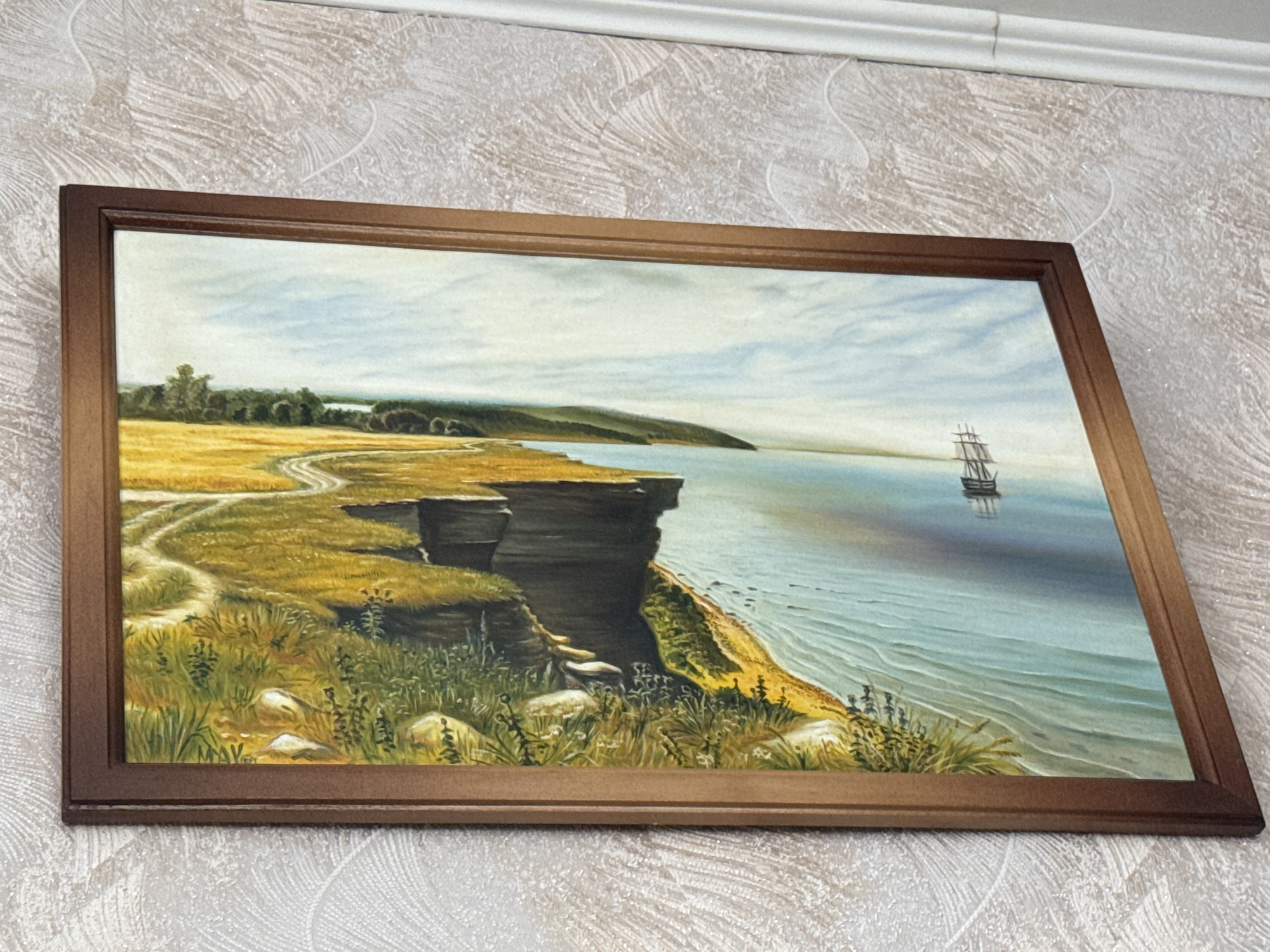 A framed oil painting depicts a seascape with a cliff overlooking a body of water. A sailboat is visible on the water. The land features a grassy cliff with a path winding through it. The sky is partly cloudy. The painting is hanging on a textured, light-colored wall. The location of this image is stated to be in the city of Seleznivka, Ukraine.