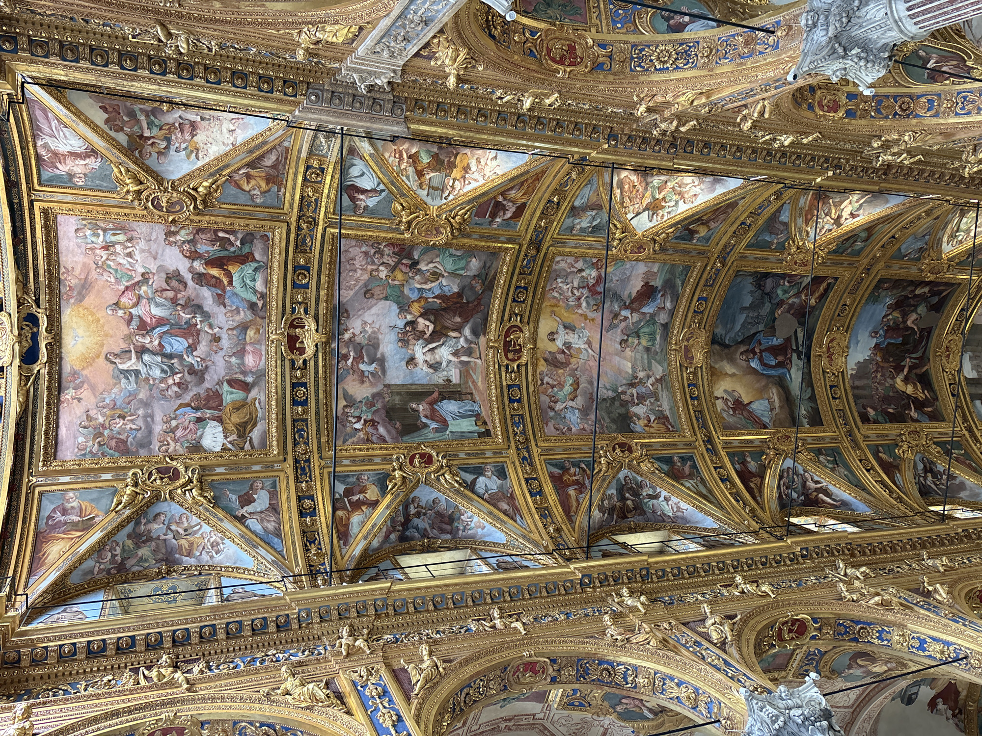 This image captures a highly ornate and gilded ceiling, likely from a grand hall or church in Genoa, Italy. The ceiling is richly adorned with numerous frescoed panels depicting intricate scenes, framed by elaborate architectural elements and abundant gold leaf.
The primary subjects are the extensive ceiling frescoes, which feature a multitude of human figures, angels, putti, and possibly saints or allegorical characters. The figures are rendered in a classical or Baroque style, often draped in flowing robes of various colors, including blues, reds, greens, and ochres. Many scenes appear to depict religious or mythological narratives, with groupings of figures interacting, ascending, or in contemplative poses. In some panels, individuals are seen reading books, while others show celestial gatherings or musical performances.
The frescoes are set within a complex structure of coffers, arches, and panels, all outlined and embellished with lavish gold gilding. This gilding forms intricate patterns, cornices, and relief sculptures, featuring delicate foliate motifs, small figures, and decorative medallions, often incorporating accents of dark blue and reddish-brown. The overall impression is one of immense grandeur and artistic mastery. Visible architectural details include a white, sculptural element in the upper right, possibly part of a column capital or a larger relief. There is no discernible text within the image. The scene conveys a sense of awe and historical richness, characteristic of significant Baroque or Rococo interiors found in cities like Genoa.