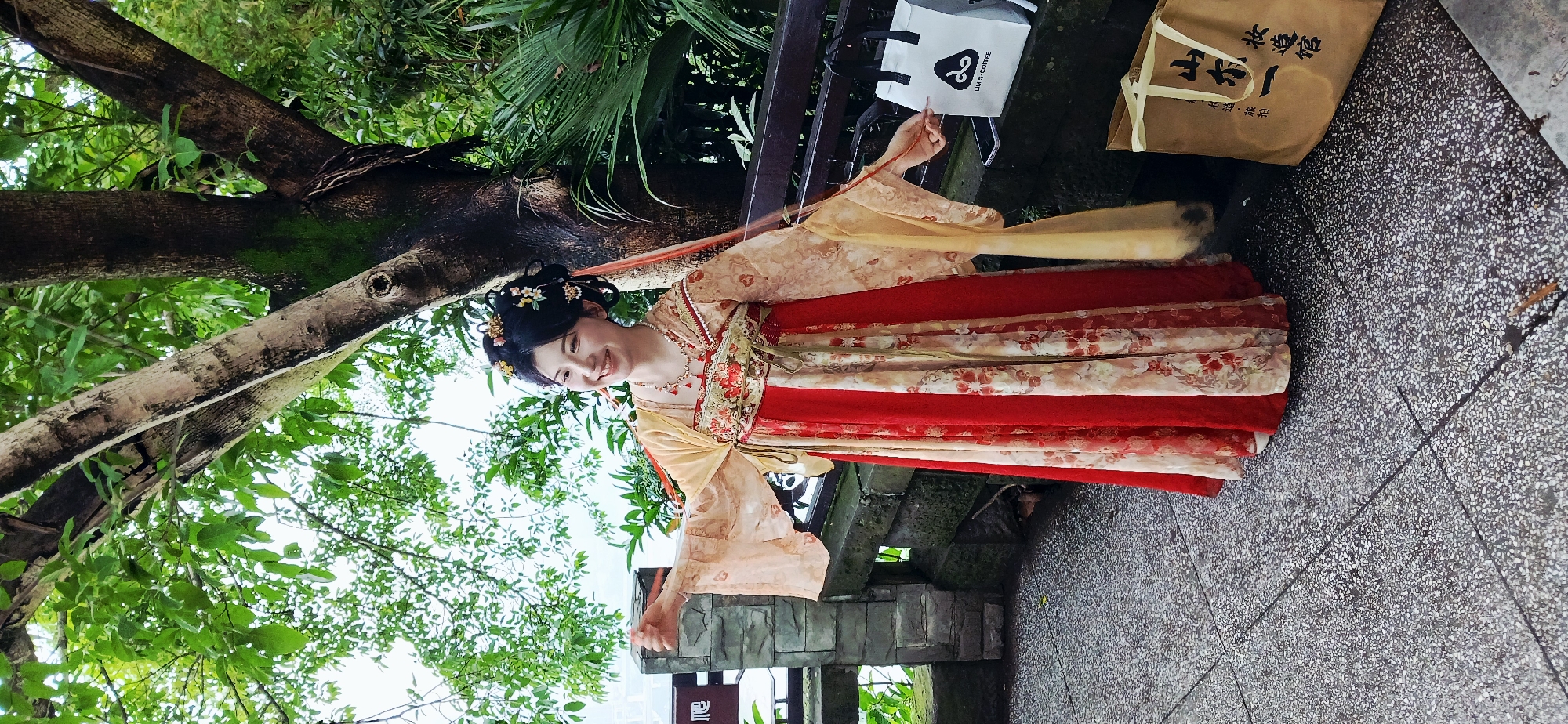 A full shot image, taken outdoors, features a woman in a traditional Chinese dress standing next to a stone structure. The woman is wearing a multi-layered dress with a red skirt and beige top with floral patterns. She has dark hair styled with flowers. The stone structure appears to be part of a walkway or overlook, with greenery visible in the background. There are two bags hanging on a railing behind her, one white with a black logo and one brown with Chinese characters. The location is the Yuzhong District in China.