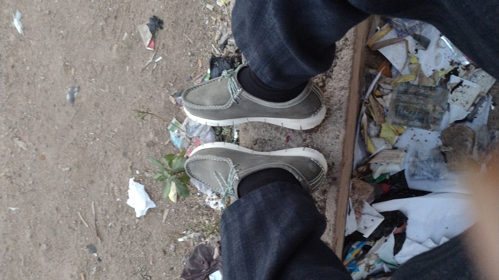 The image shows a person's lower body from a high angle. The person is wearing gray-green moccasin-style shoes with white soles and light green laces, black socks, and dark blue denim jeans. The person is standing in a cluttered outdoor environment. To the left of the person is dirt. To the right of the person, inside what looks like a shallow wooden frame or box, is an accumulation of trash. The location may be Jos, Nigeria, as requested.