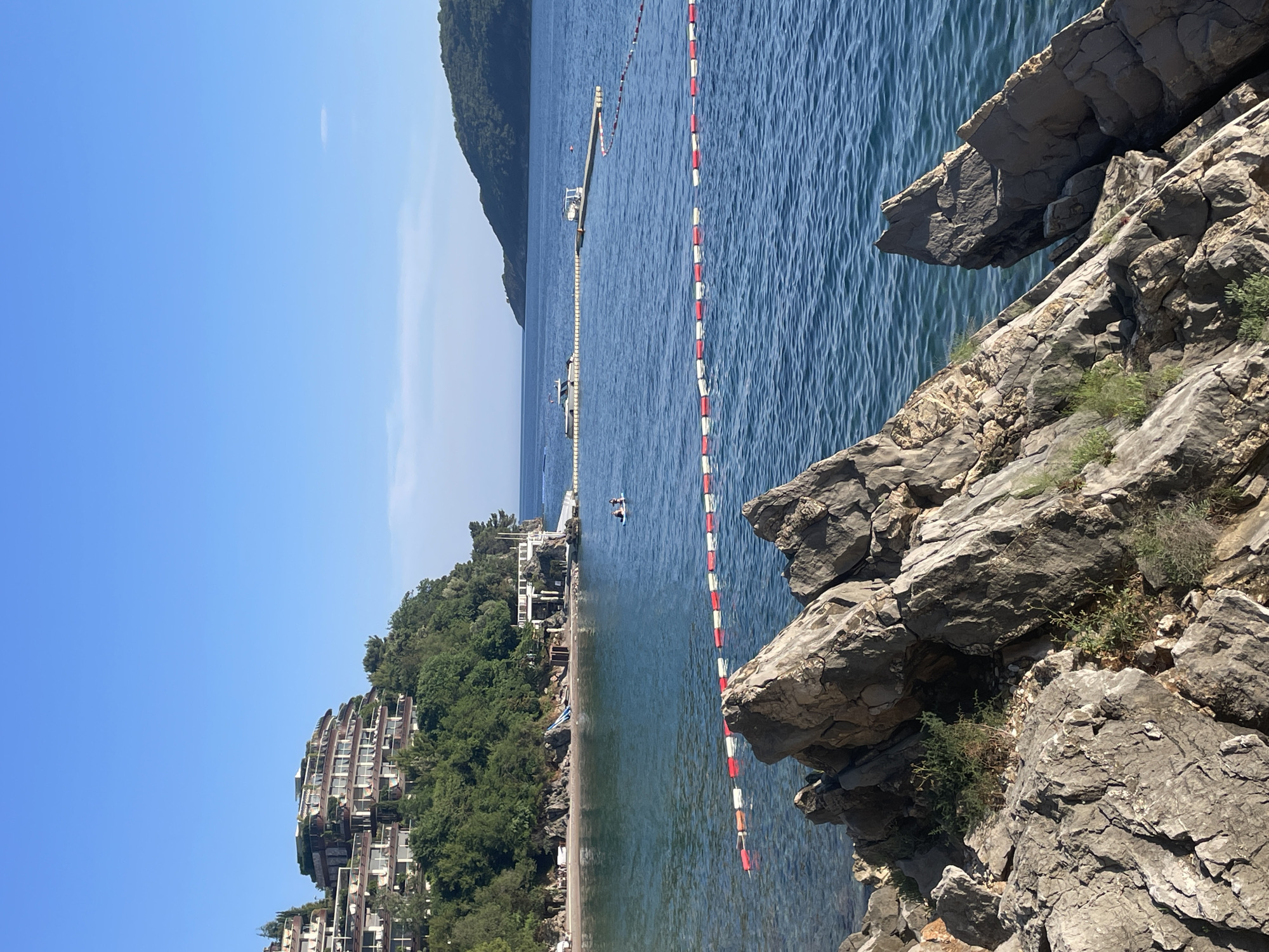 The image shows a view of a coastline in Budva, Montenegro. A body of water is partially visible, divided by a red and white safety rope. On the left side of the frame, a beach is lined with buildings and green trees. In the distance, an island is seen. In the lower right corner, there are large, gray rocks. A person is paddling in the water. The sky is clear and blue.