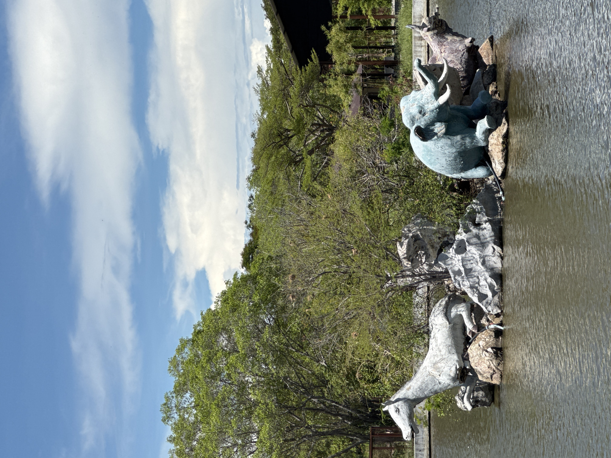 This outdoor shot in Bang Pu Subdistrict Municipality, Thailand, features animal sculptures within a body of water. The sculpture on the left appears to depict a horse or pony, while an elephant sculpture sits towards the upper right of the frame. Both figures are set amongst rocks and vegetation. The water surface shows subtle ripples. Behind the sculptures are trees and foliage, with a partial view of what appears to be a building or structure. Above, a blue sky is visible with scattered white clouds.