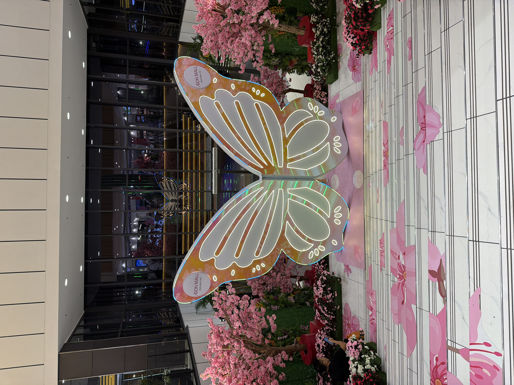 The image depicts a festive mall interior, likely decorated for a seasonal event. Dominating the center is a large, illuminated butterfly sculpture with intricate white outlines. To the left and right of the sculpture are artificial trees with abundant pink blossoms, resembling cherry blossoms. The foreground features a set of stairs decorated with painted pink flowers and floral motifs on a white tiled surface. A person is seated on the stairs, using a mobile phone. In the background, large glass windows reflect the mall's interior, showing escalators and other shoppers. The overall impression is one of a vibrant, decorated public space. The specific location of An Cựu Ward, Vietnam, is inferred based on the subject matter and visual cues, though not explicitly stated in the image itself.