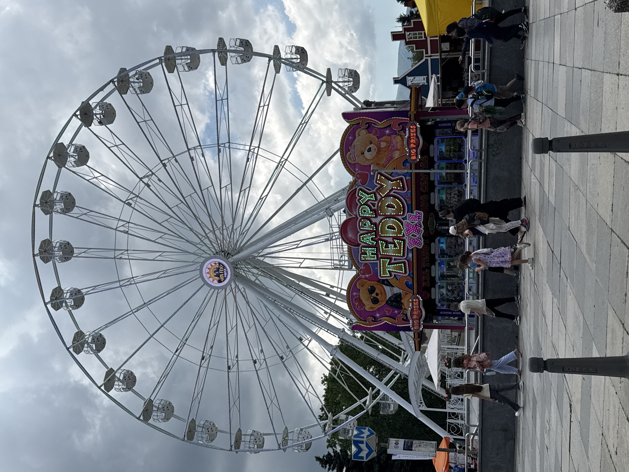 An outdoor scene captures a bustling fairground or amusement park, dominated by a large white Ferris wheel on the left. The sky is overcast with significant cloud cover, suggesting a cloudy day. Several people are visible on the paved ground, walking or standing near various attractions.
The Ferris wheel features numerous cabins and its central hub displays an illuminated sign that reads "SUNFEST SOFIA." To the right of the Ferris wheel, a colorful game booth or attraction is visible, adorned with bright lights and signs. Visible text on these signs includes "HAPPY TEDDY," "BIG PRIZES" (repeated), and "XXL TEDDY," indicating a teddy bear themed game. Other partial texts like "WINNING CENTRE" and "WINDING WHEEL" are also discernible. Further to the right, behind the game booths, are colorful buildings (red, blue, yellow) and green trees. The foreground features a paved area with a checkered pattern and a dark grey bollard with partially visible text "OOSS." Below the Ferris wheel, a blue sign with "MM" is seen. The overall atmosphere is that of a lively public event or carnival in Sofia, Bulgaria.