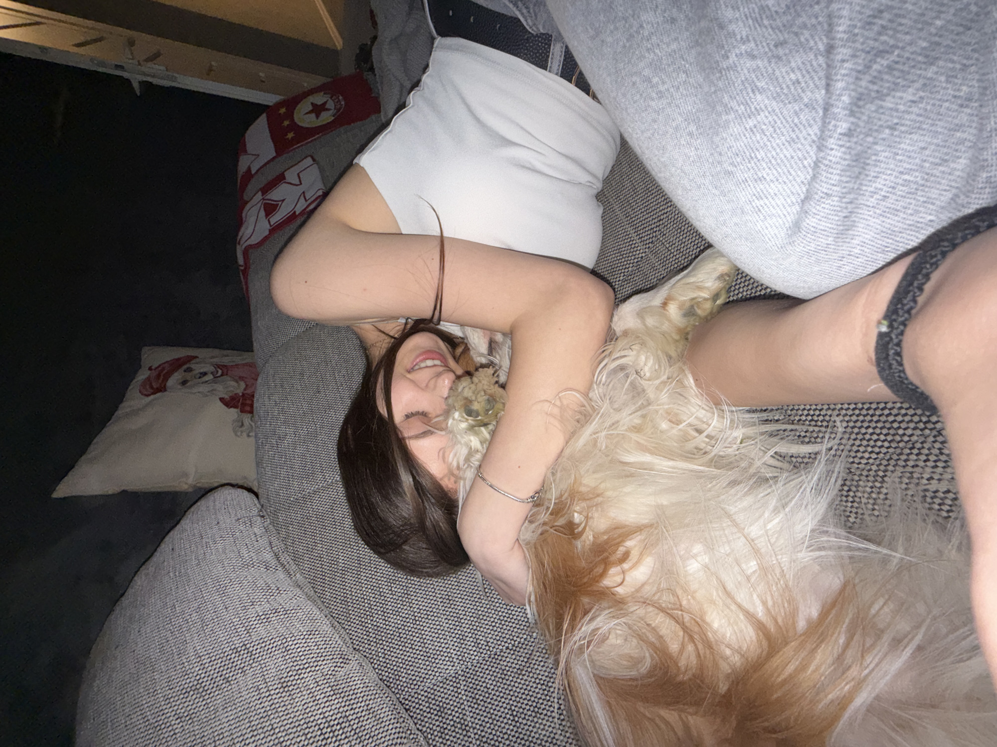 A woman is lying on a gray patterned sofa, facing the camera and smiling. Her head is positioned towards the left side of the image. A white and tan long-haired dog is snuggled next to her, with its paw near her face. The woman is wearing a white top and gray pants, secured with a black belt. A patterned cushion is visible in the background on the left side. Part of a shelf is visible in the upper left corner. The woman is wearing a bracelet on her left wrist and a black band around her right leg. The location is likely indoors. Information regarding the city of Sofia, Bulgaria, is not discernable from the image.