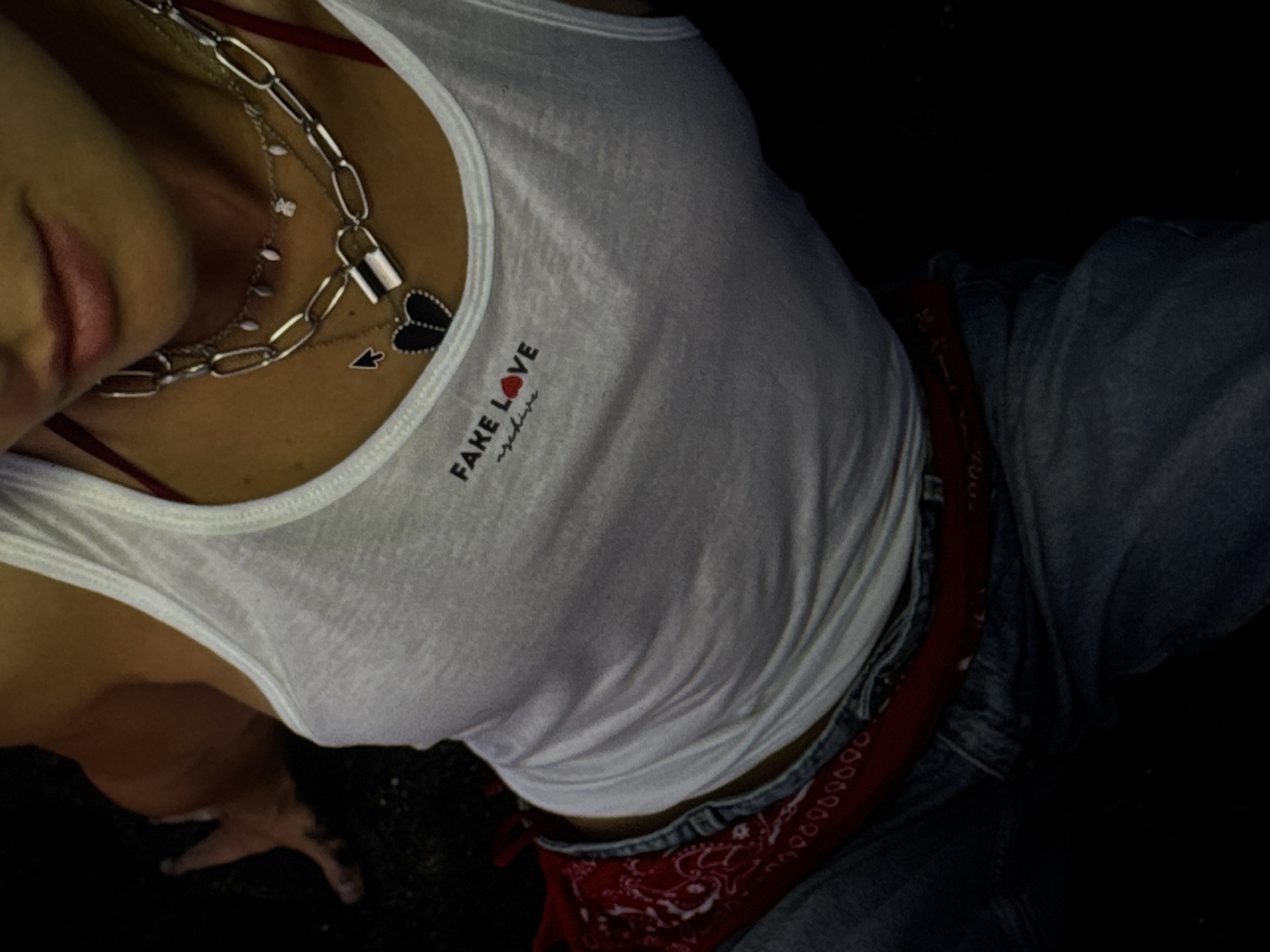 An individual is seated, wearing a white tank top with the text "FAKE LOVE archive" and a red heart graphic. They are also wearing denim jeans and a red bandana is visible around their waist. The individual is adorned with several necklaces, including one with a padlock pendant and another with a black heart pendant. The setting appears to be indoors or dimly lit, with a dark background. There are no explicit indicators of the location being Kuta Selatan, Indonesia.