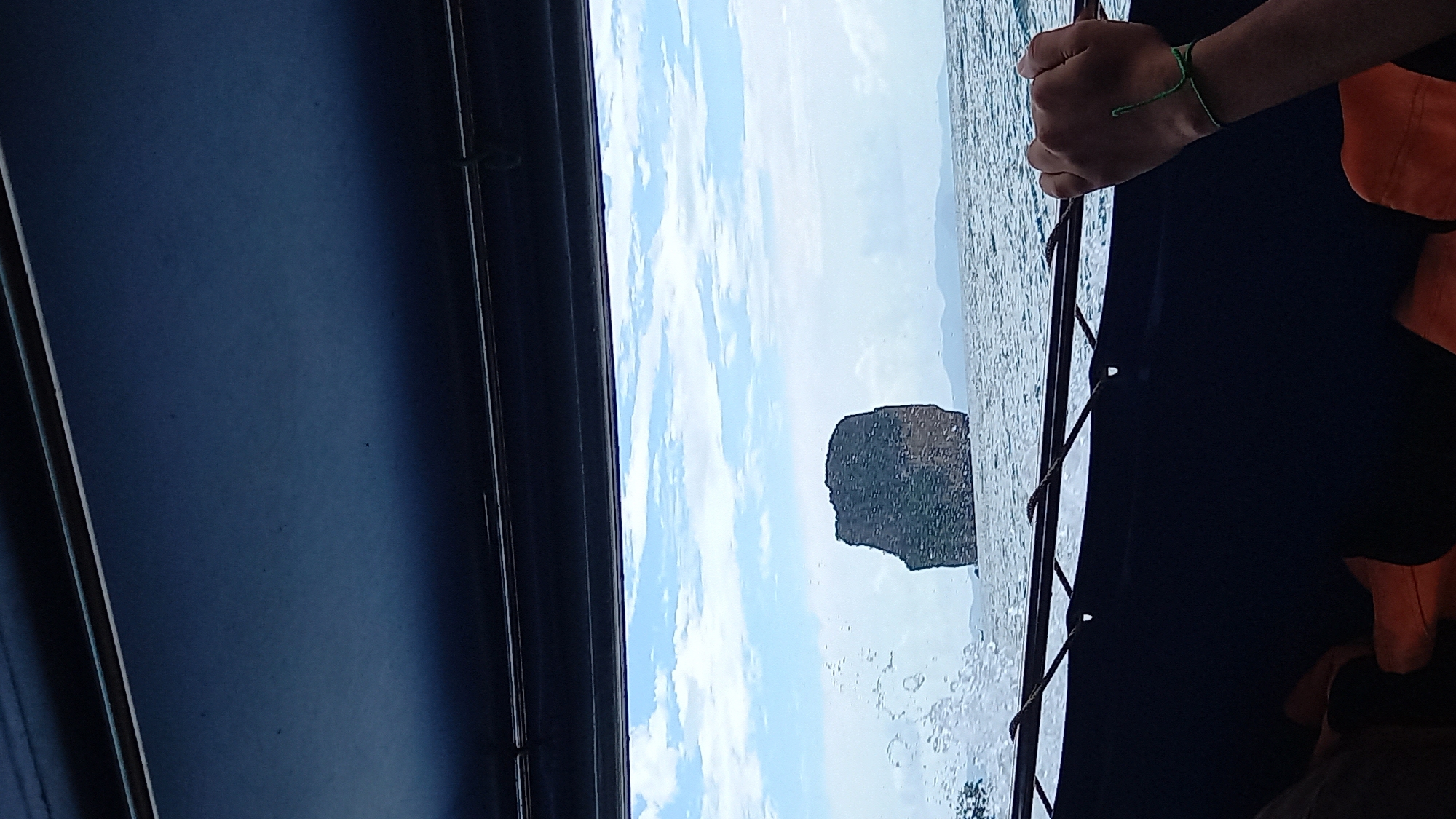 The view from inside a boat in Ao Nang, Thailand, shows a rocky island in the sea under a cloudy sky. The dark interior of the boat frame the outdoor scenery. A person's hand, adorned with a green bracelet, grips a rope attached to the boat's frame. A life vest is visible on the person wearing the vest. The boat appears to be moving, indicated by water droplets on the window.