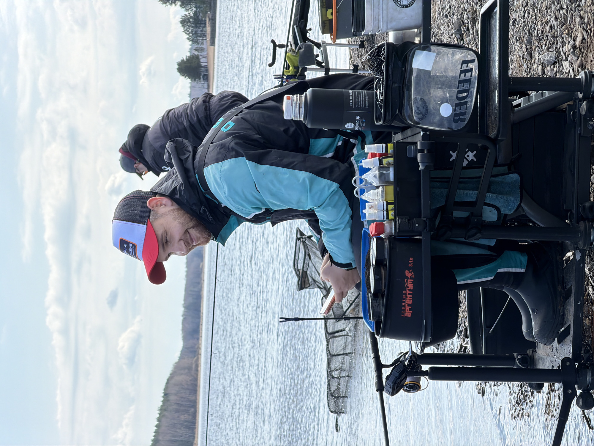 Two men are engaged in feeder fishing on the rocky bank of a large lake or reservoir, likely in Chaikovsky, Russia. The man in the foreground, wearing a blue and black jacket and a red and white baseball cap (with partially visible text "ARCENS"), is leaning forward, focused on his extensive fishing setup. He appears to be preparing bait or adjusting his gear, displaying a slight, content smile. Another individual in a dark jacket is partially visible behind him.
The scene is dominated by specialized fishing equipment. A sturdy black fishing station holds various accessories, including a clear zippered pouch labeled "FEEDER," a black insulated bottle with "HEALTH. SAFETY." and "CARBON CUP 798" printed on it, and several small containers with liquids or bait. A black bucket or container visible on the station is labeled "FISHING АРГЕНТУМ 3.1л" (Argentum 3.1L). Fishing rods extend towards the water, and a landing net is partially submerged. A white bucket with partially visible text "CHAMPI..." can be seen in the background.
The weather appears cool, as both men are dressed in jackets. The sky is partly cloudy, indicating daytime. The background features a vast body of water, with a forested shoreline visible in the distance. The immediate ground is covered with pebbles and small rocks.