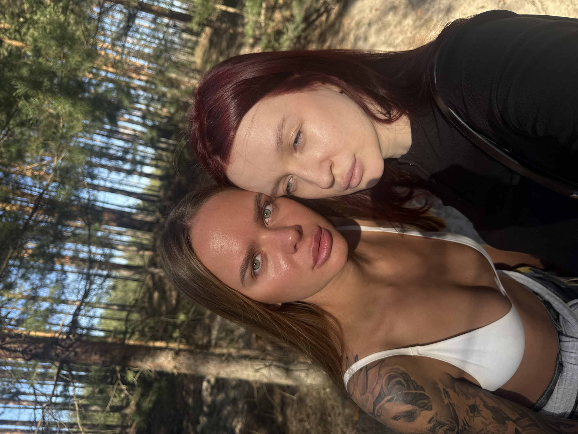 Two women are posing for a photograph outdoors in a forest-like environment, possibly near Nizhny Novgorod, Russia. The woman in the foreground has light brown hair and a tattoo on her left shoulder; she is wearing a white top. The woman behind her has red hair and is wearing a black top. They are standing close together, with the red-haired woman resting her head on the other woman's head. The background shows trees and sunlight.