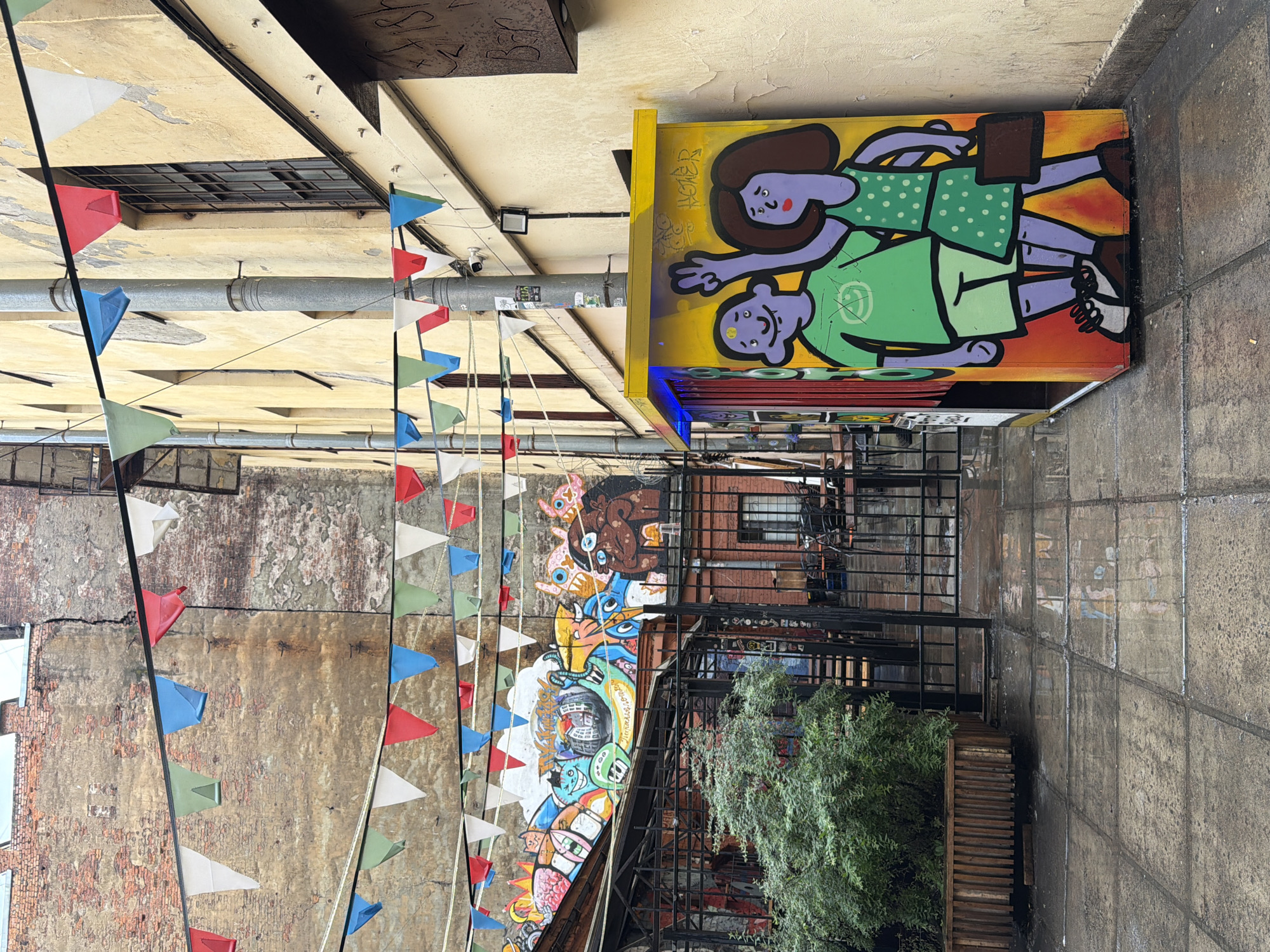 The image depicts an outdoor urban courtyard. On the right, a bright yellow kiosk is adorned with graffiti art of two purple figures, a male in a green t-shirt and shorts, and a female in a green polka-dot dress, both waving. The kiosk has blue lighting at the top. The ground is paved with large square tiles, wet and reflective from recent rain, with scattered cigarette butts. To the left, a large wooden planter box contains a lush green bush. Beyond the planter, a black metal gate leads into a covered area with tables and chairs, suggesting an outdoor seating space for a cafe or restaurant. Above the gate, a vibrant mural covers the wall, featuring a colorful array of cartoonish figures and abstract designs. Decorative triangular flags in red, blue, white, and green are strung across the courtyard, attached to the buildings on either side. The buildings themselves are old, with textured brickwork and peeling paint. A large metal pipe runs vertically along the right-hand building. The overall atmosphere is that of a lively, artistic, and somewhat gritty urban space.