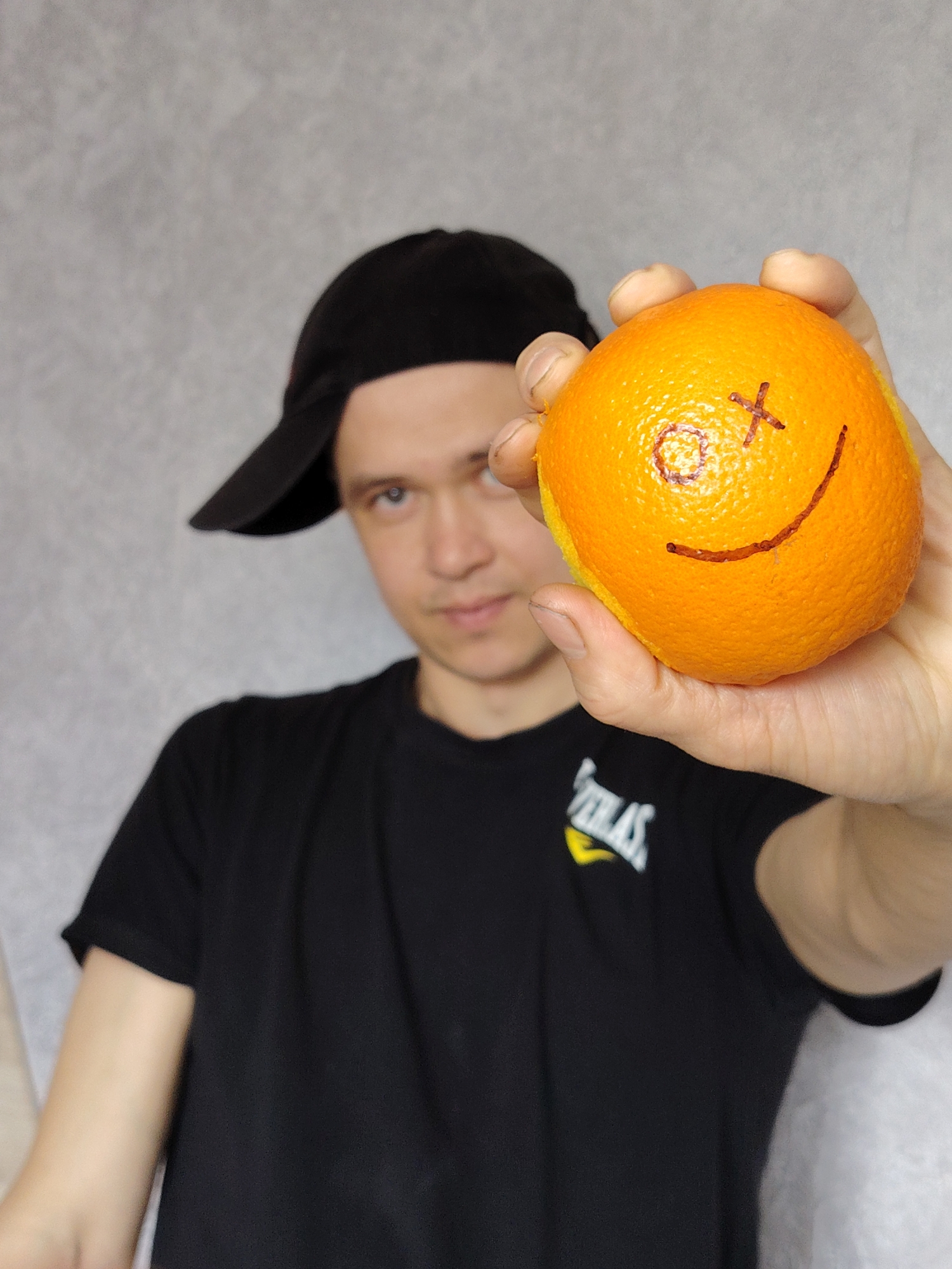 A young man wearing a black baseball cap and a black t-shirt stands against a gray background, holding an orange with a drawn face in his outstretched hand. The face on the orange has crossed-out eyes and a curved line for a smile. The location is the city of Chaikovsky, Russia.
