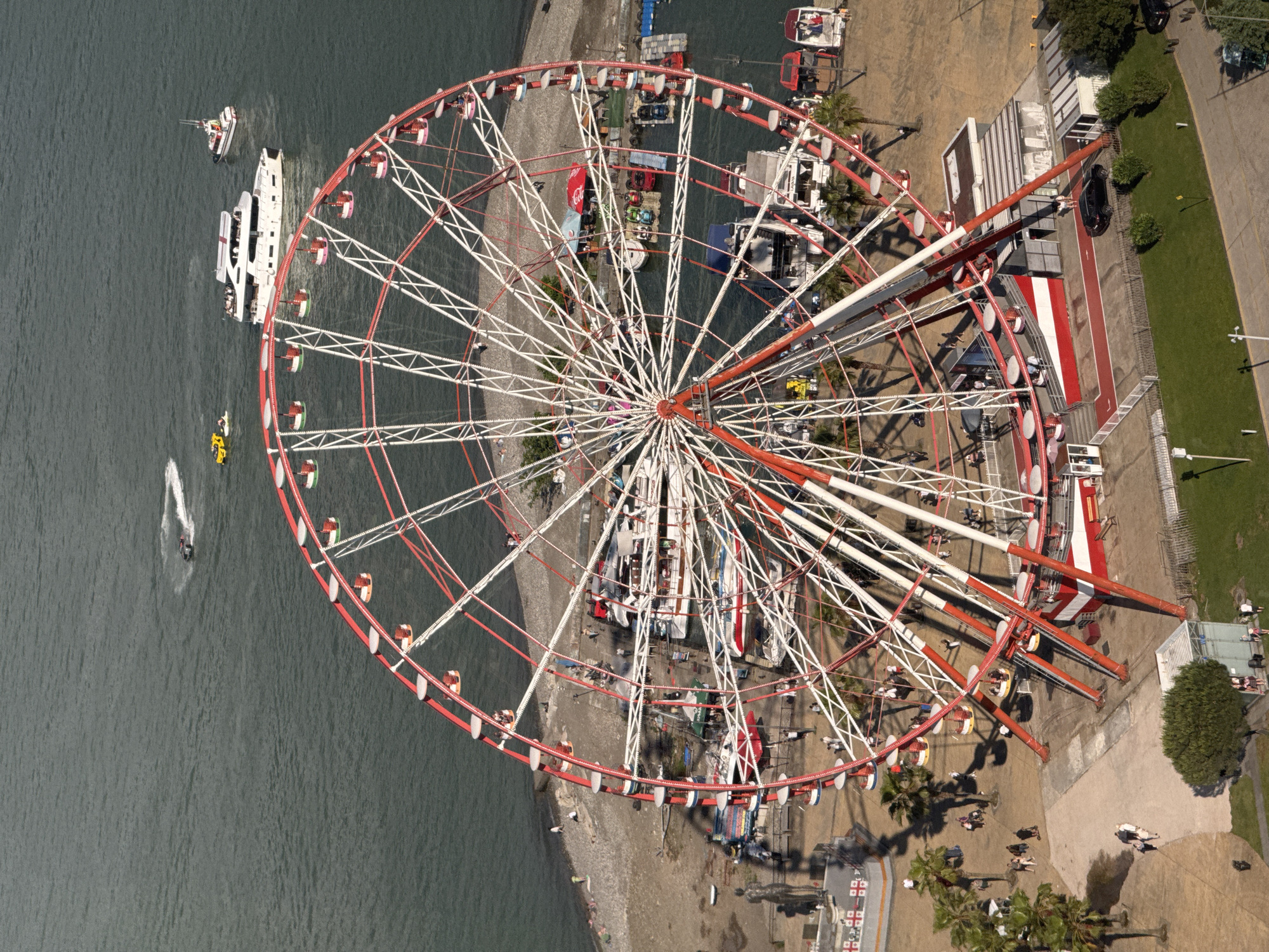 This aerial photograph captures a large Ferris wheel on a sunny day, likely late morning or early afternoon given the shadows. The Ferris wheel, a prominent red and white structure, dominates the frame. Its numerous gondolas, painted in various colors including red, yellow, and green, are visible as they ascend and descend.
The Ferris wheel is situated near the coastline, with a body of water, likely the Black Sea, to its left and a beach area along its base. Several boats are present in the water: a white passenger boat with multiple decks, a smaller white boat, and a yellow jet ski creating a white wake. People can be seen on the beach and on the boardwalk area surrounding the Ferris wheel, some appearing to be walking or gathering.
Beyond the beach, there's a paved area with palm trees and more people. A red and white striped platform, possibly part of the amusement park infrastructure, is attached to the Ferris wheel's base. Further to the right, there is a grassy area and a paved road, suggesting a developed promenade or park.
The scene appears to be a lively tourist area, with the Ferris wheel serving as a central attraction. The weather is clear and bright, indicated by the strong sunlight and distinct shadows. The location cues, such as the architecture of the Ferris wheel's support structure and the presence of palm trees, are consistent with a coastal resort town. No discernible text is visible in the image.