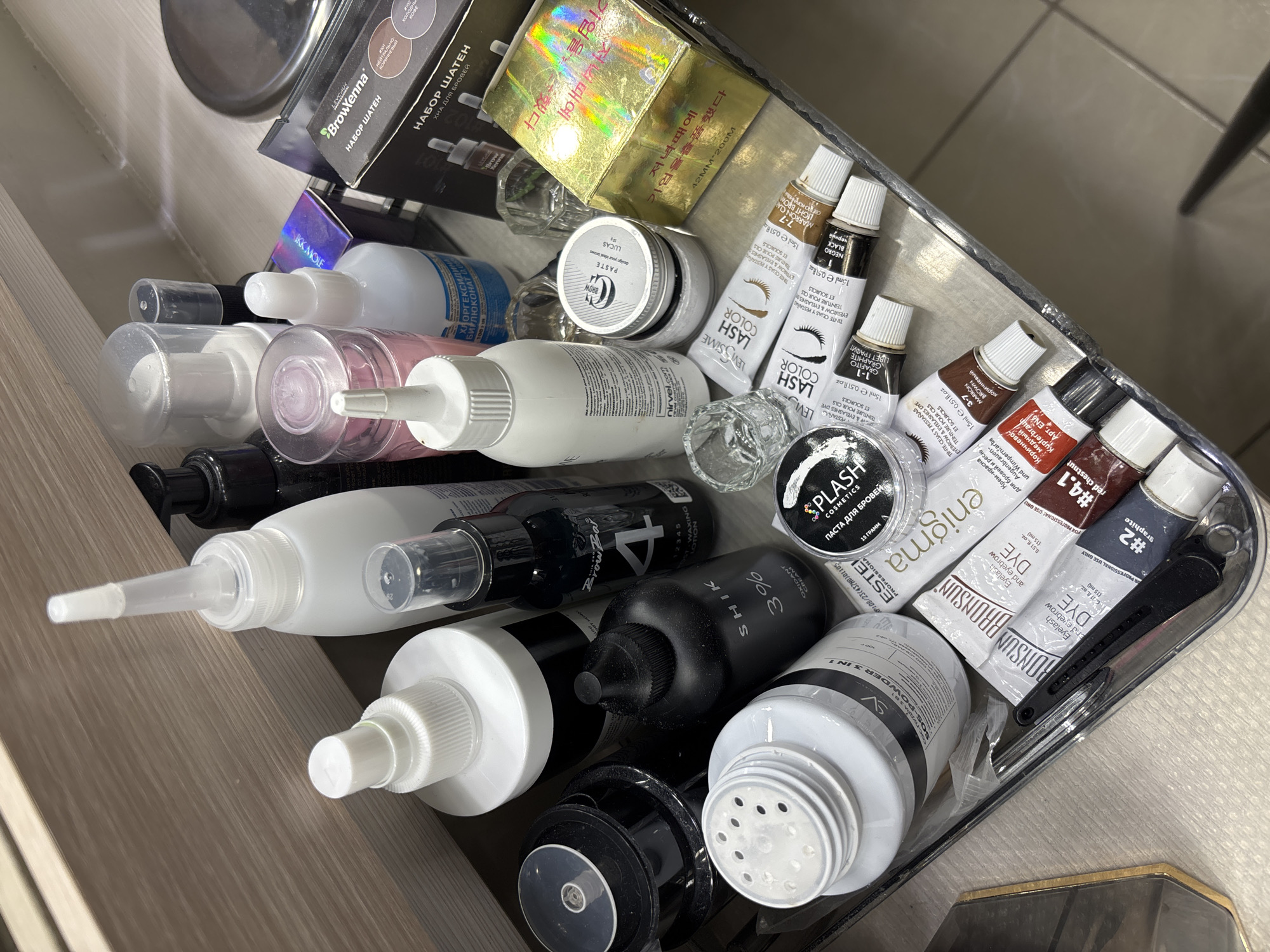 This is a close-up, top-down shot of a collection of beauty products, primarily for eyebrows and eyelashes, neatly arranged in a drawer or container. The items are predominantly small bottles, tubes, and jars. Among the visible products are several tubes of "LASH COLOR" and "EYEBROW & EYELASH DYE" from brands like LEVISSIME and BRONSUN, indicating a focus on tinting. There are also bottles of "BrowXenna" eyebrow henna kit, "SHIK 3% OXIDANT CREAM", and "PLASH COSMETICS" eyebrow paste. Other items include what appears to be a liquid labeled "БИЛЮКОНАТ КЛОРГЕКСИДИН", suggesting a disinfectant or cleaning solution. The overall impression is that of a professional beauty kit or a personal collection of cosmetic supplies. The lighting is consistent and suggests an indoor setting, likely a beauty salon or a personal vanity area. There are no people or pets visible in the image. The scene is well-lit, highlighting the various packaging and labels of the products. The visible text includes product names, brand names, ingredient information, and usage instructions in multiple languages, including Russian, English, Spanish, French, and Korean.
