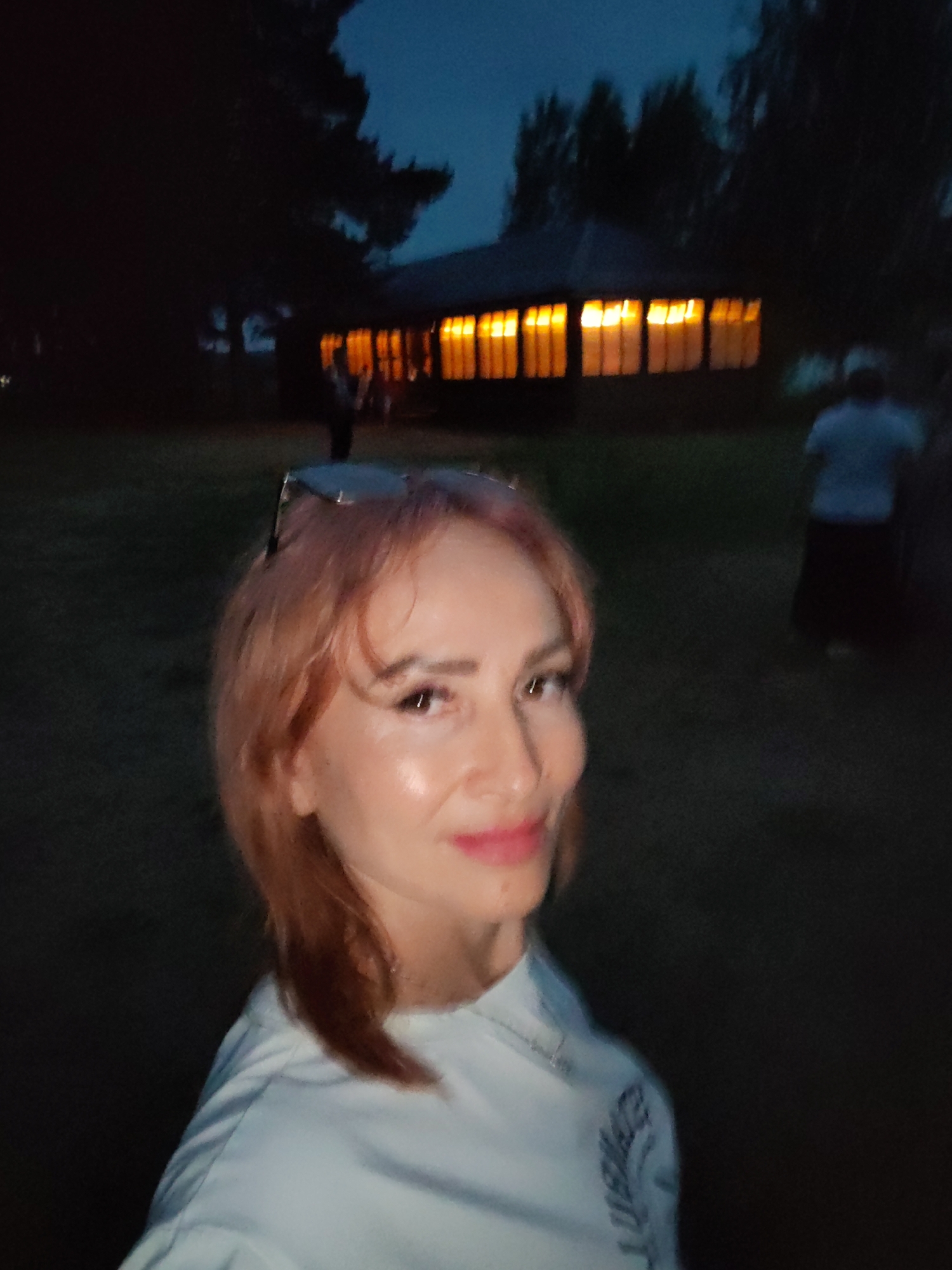In the foreground, a woman with reddish-blonde hair and pink lipstick is captured in a close-up selfie. She is wearing a light blue sweatshirt. Sunglasses are perched on top of her head. In the blurred background, a building with illuminated windows is visible, suggesting it is evening or night. Trees surround the building and the general area. The scene appears to be outdoors in a natural or park-like setting, possibly within the city of Chaikovsky, Russia, given the prompt. Two other individuals are faintly visible in the background, one near the building and another further away.