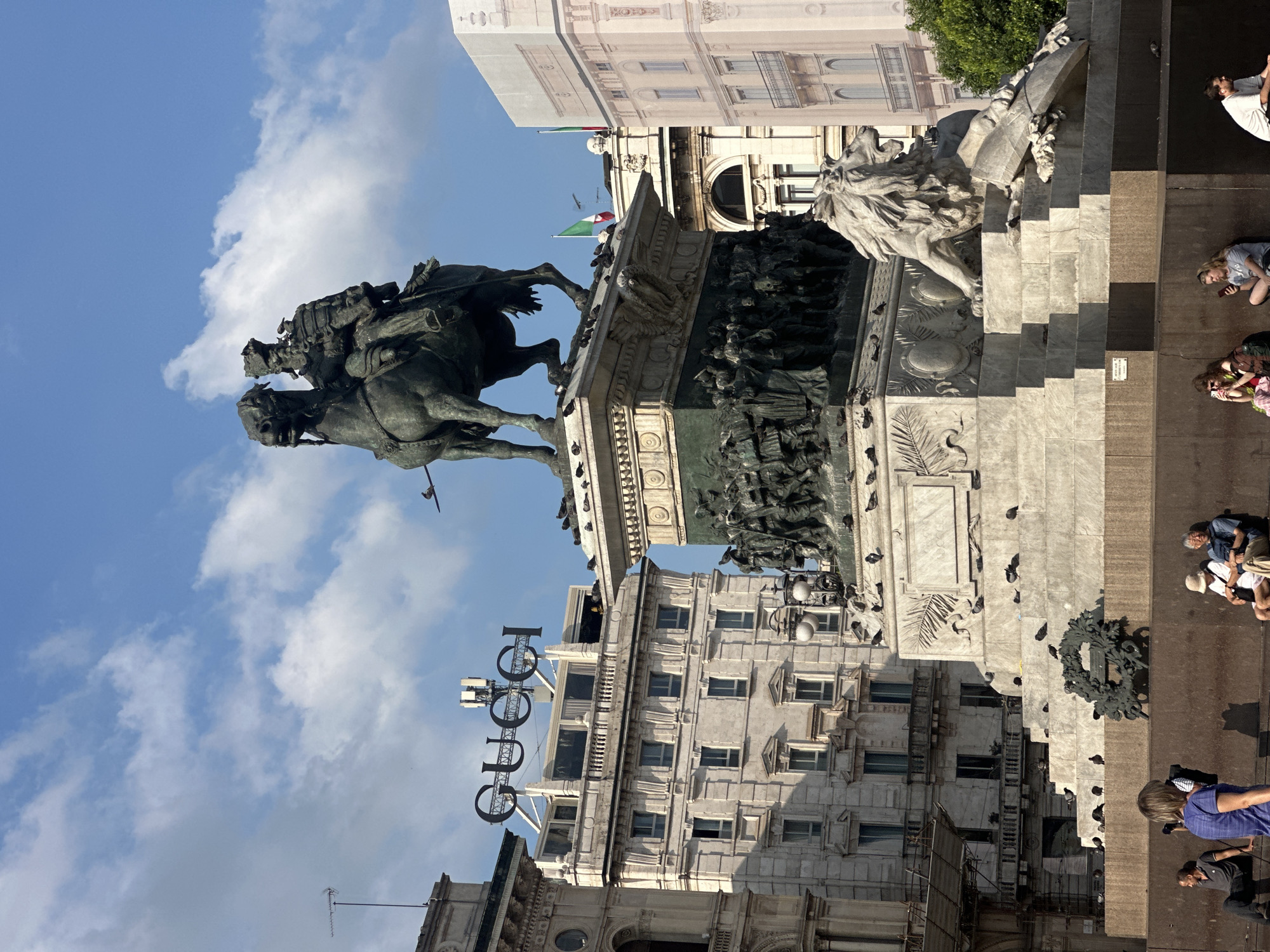 The image depicts the equestrian statue of Victor Emmanuel II in Piazza del Duomo, Milan, Italy. A bronze statue of the king on horseback stands prominently atop a large marble pedestal adorned with relief sculptures and winged figures. To the right of the pedestal, a broken marble lion sculpture is partially visible. The statue and pedestal are populated by numerous pigeons. In the background, several buildings are visible, including a light-colored building with the prominent "GUCCI" logo on its facade, and other classical European architecture. Italian flags are flown from some of the buildings. In the foreground, several people are seated and standing. One person in the foreground is holding a camera. The sky is blue with scattered white clouds.
