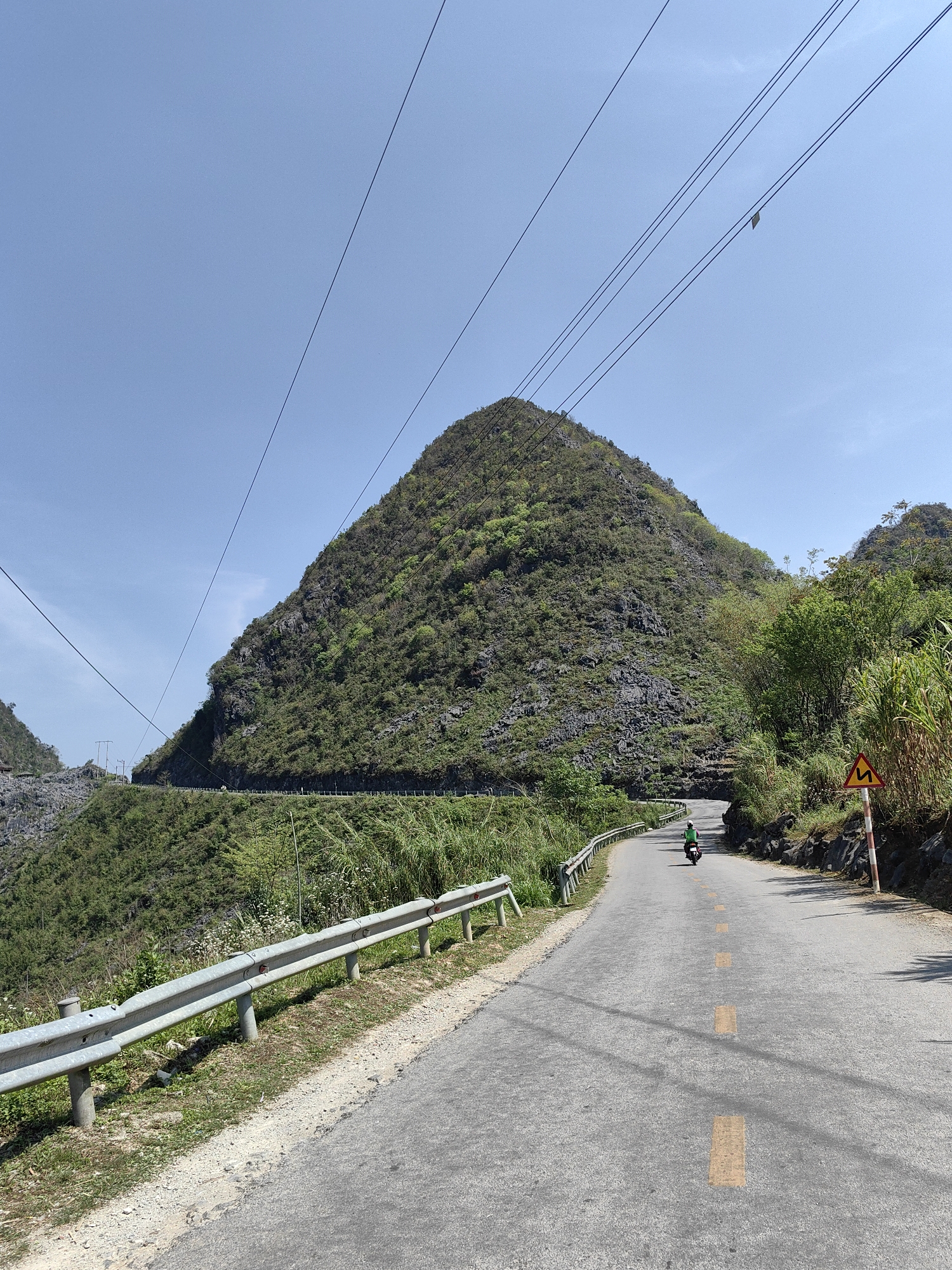 The image shows a paved road winding through a mountainous landscape in Sà Phìn Commune, Vietnam. A motorcyclist is traveling along the road, which is marked with yellow lines. A guardrail runs along the left side of the road. A yellow and red road sign indicating a curve is visible on the right side. A large, pyramid-shaped mountain covered with vegetation dominates the background. Power lines stretch across the sky. The sky is clear and blue.