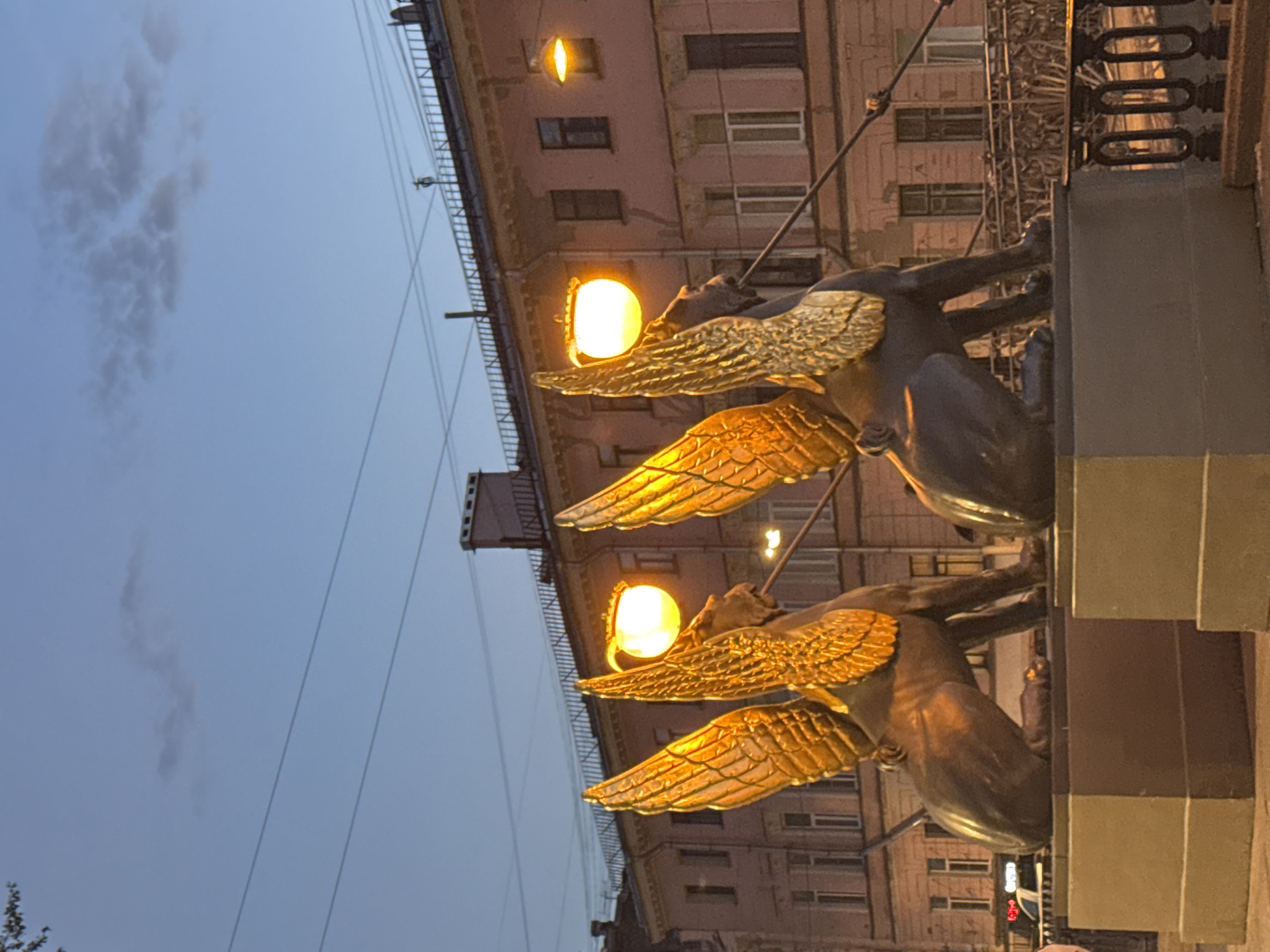 The image shows two winged lion statues, known as griffins, on the Bank Bridge in Saint Petersburg, Russia. The griffins are made of bronze and have golden wings, with their heads facing forward. Each griffin is illuminated by a globe-shaped lantern on a pole extending from its back, casting a warm glow. Behind the statues is a building with a pink facade, featuring numerous windows and a decorative railing along the roofline. The sky above is a pale blue with some wispy clouds. The griffins are situated on pedestals, and the scene appears to be a street or bridge setting, as indicated by the railing and the presence of a road or walkway.