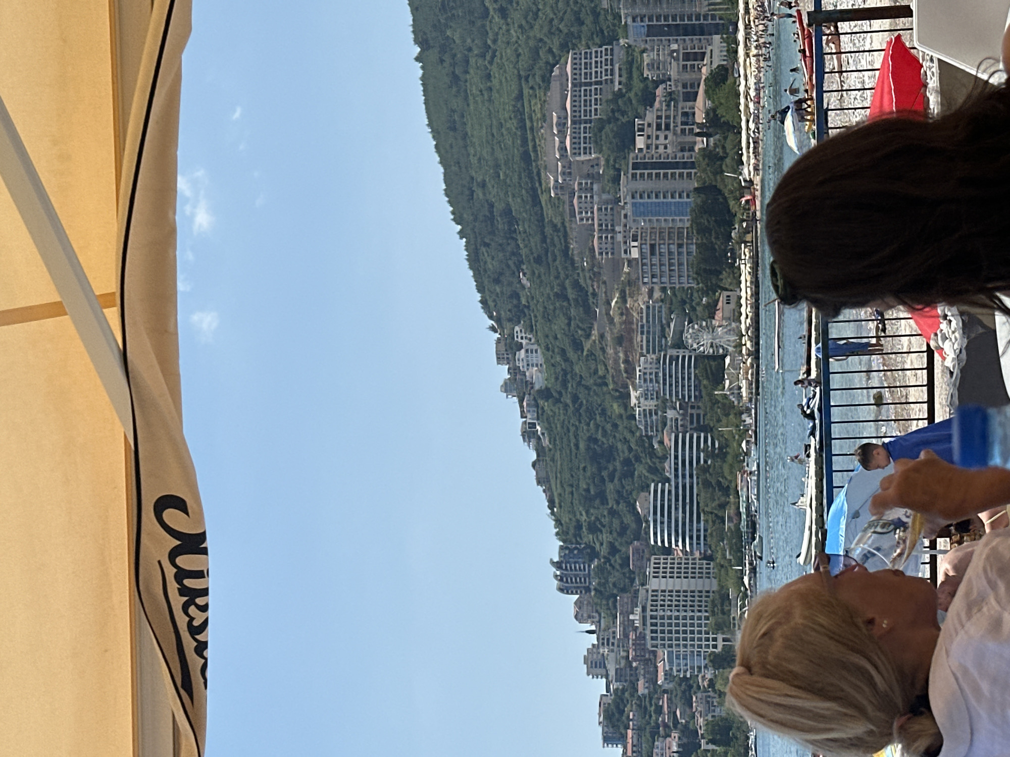 In Budva, Montenegro, a woman with blonde hair pulled back in a ponytail is seated at an outdoor cafe, holding a glass of a light-colored beverage to her lips. To her right, the back of a woman with dark, long hair is visible. They are positioned under a beige awning with black lettering on the underside. In the foreground, a blue water bottle is partially visible. Beyond them, a metal railing separates the seating area from a pebbled beach. Several people are on the beach, with some in the water. A child in a blue towel stands near the railing. Further in the background, boats are anchored in the calm sea. The scene overlooks a coastline lined with numerous hotels and buildings ascending a densely vegetated hillside. The sky is clear and blue with a few scattered white clouds.