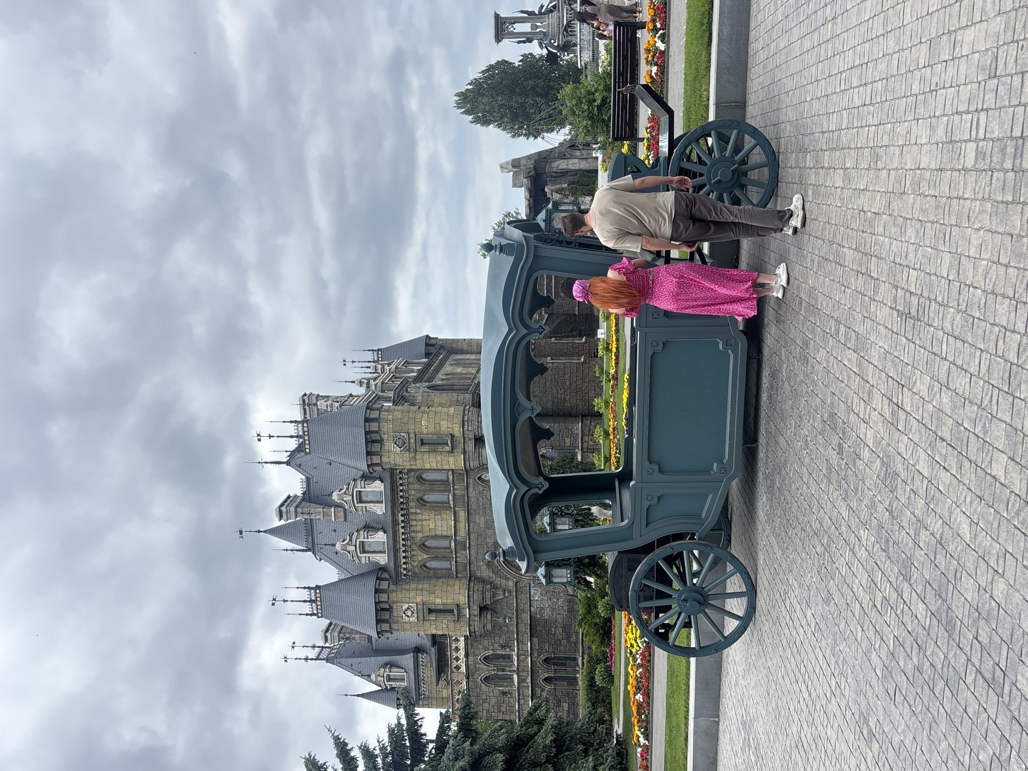 In Khryashchevka, Russia, two people stand near a decorative carriage in front of a castle-like building. The man, wearing a tan t-shirt and grey pants, is on the right, his back mostly to the camera. A woman with bright red hair, wearing a pink polka-dot dress and a matching headscarf, is to his left, facing the carriage. They appear to be interacting with or observing the carriage.
The carriage is dark teal with spoked wheels and ornate detailing. It is positioned on a paved walkway. Behind the carriage and people, a large, multi-turreted building with grey roofs and stone walls dominates the background, resembling a fantasy castle. Flowerbeds with colorful blooms of red, yellow, and white line the path in front of the building, with a strip of green grass adjacent to them. Trees, including evergreens, are visible to the left of the carriage and building.
The sky is overcast with heavy grey clouds, suggesting a cloudy day. The lighting is diffused, indicating it is likely daytime, but the specific time is not discernible. The overall scene evokes a sense of being at a tourist attraction or theme park, given the elaborate architecture and decorative elements. No visible text is present in the image.