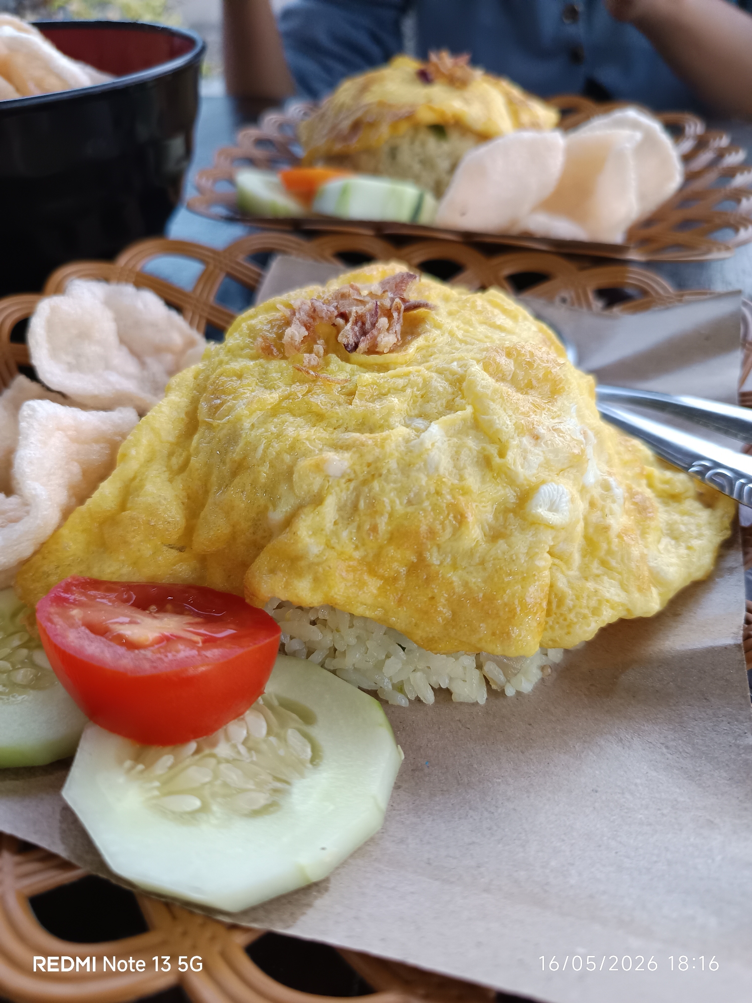 A close-up shot shows a plate of food served on a woven brown tray. The main dish appears to be rice covered with a large, yellow omelet, garnished with fried onions. Accompanying the omelet are several slices of cucumber, half a tomato, and some crackers. In the background, another plate of the same dish is visible, along with a black bowl. The scene appears to be set outdoors, possibly at a restaurant or cafe in Tanjung Morawa, Indonesia, based on the "REDMI Note 13 5G" watermark at the bottom left and the timestamp "16/05/2026 18:16" at the bottom right of the image, indicating the photo was taken on May 16, 2026. A person is partially visible behind the second plate of food, wearing a blue shirt. Utensils are resting on the plate near the main dish. The food is placed on a sheet of plain brown paper.