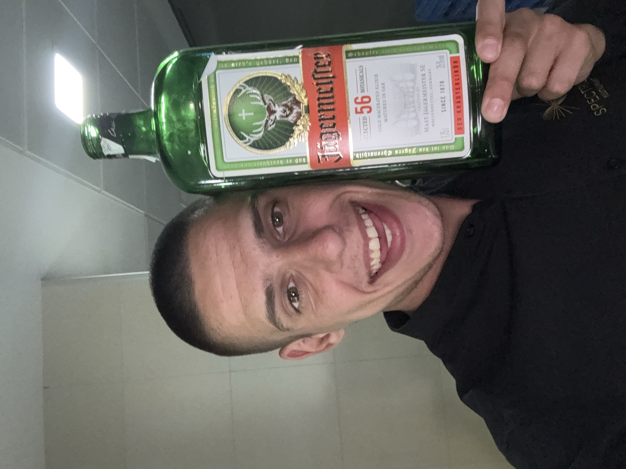 A man with short, dark hair is posing for a selfie with a bottle of Jägermeister balanced on his head. He is smiling broadly, showing his teeth. The bottle is green glass with a distinctive label featuring a deer head emblem. The man is wearing a black shirt, and the logo of "SPECTRUM" is visible on his shirt. The background appears to be an indoor space with plain walls and overhead lighting. There is no evidence in the photograph to pinpoint a location in Sofia, Bulgaria.