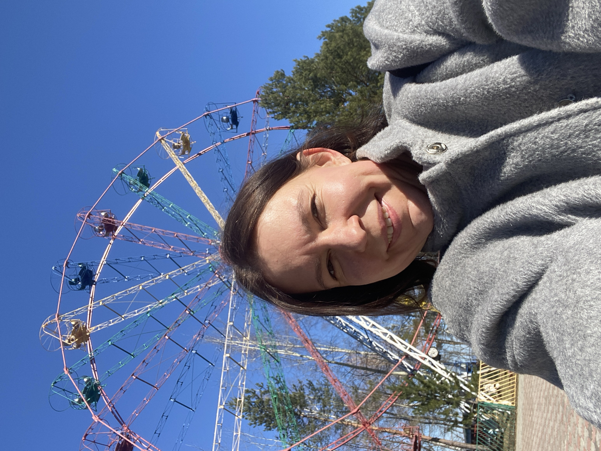 A woman is standing in front of a Ferris wheel in Chaikovsky, Russia. The woman is facing the camera and smiling. She is wearing a gray coat. The Ferris wheel is colorful, with blue, green, red, and yellow spokes. The sky is blue and clear. There are trees in the background.