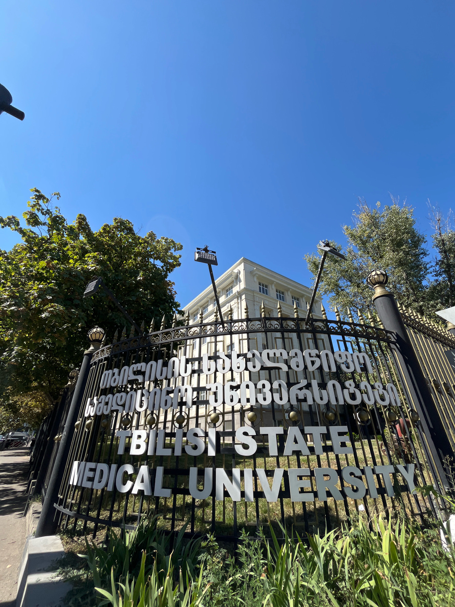 The image shows a sign in front of a building with the words "TBILISI STATE MEDICAL UNIVERSITY" in large white letters. The sign is on a black metal fence with gold accents. The building appears to be a large, light-colored, multi-story structure. Trees and foliage are visible in front of and around the building. The sky is clear and blue. The image is taken from a low angle, looking up at the sign and building.