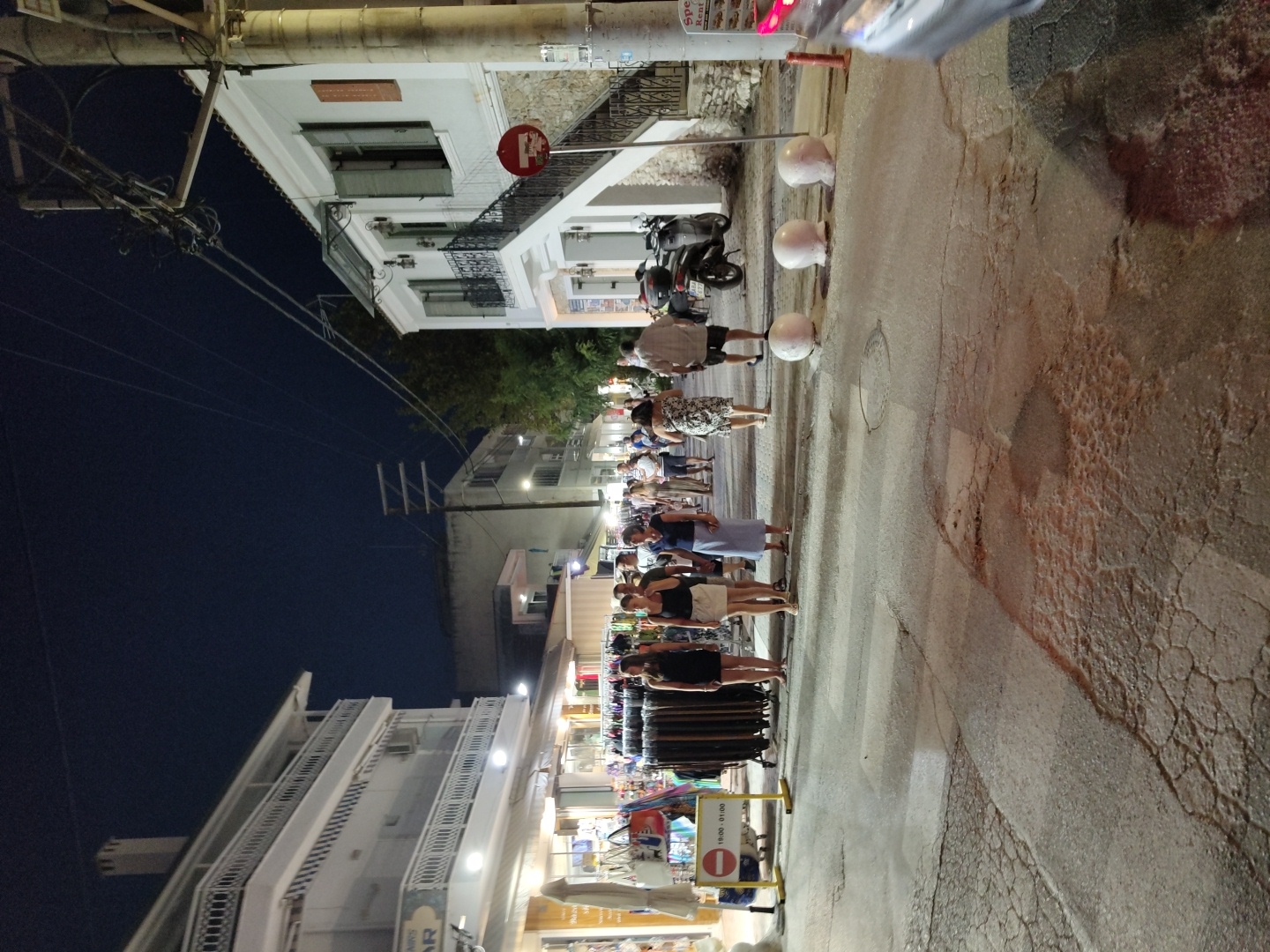 The image depicts a nighttime scene on a street in Limenaria, Greece. A group of people are walking along the street, which is lined with buildings and shops. There are also several motorcycles parked on the side of the street. The street appears to be paved with asphalt, and there are some cracks and unevenness in the surface. The buildings on either side of the street are whitewashed, and some have balconies with railings. Overhead, there are power lines and street lights, illuminating the scene. The sky is dark, indicating it is nighttime. Several white spheres are placed on the ground on the right side of the street, possibly as bollards. A red and white circular sign is visible on a pole in front of one of the buildings.