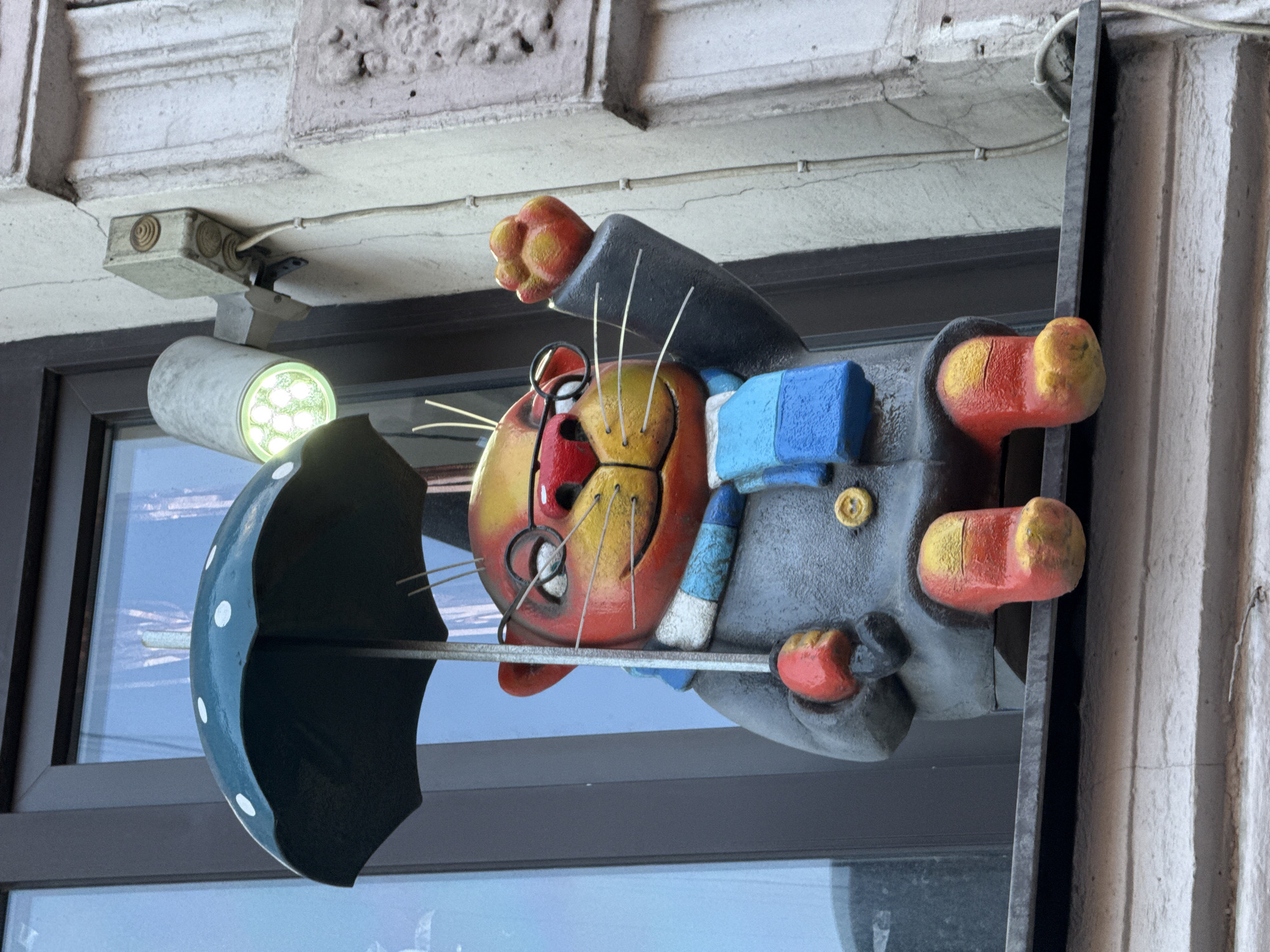 A whimsical statue of a cat wearing glasses, a coat, and a scarf sits on a windowsill. The cat is holding a polka-dotted umbrella and has one paw raised. A light fixture with green LEDs is mounted on the wall above the statue. The scene is located in Saint Petersburg, Russia.