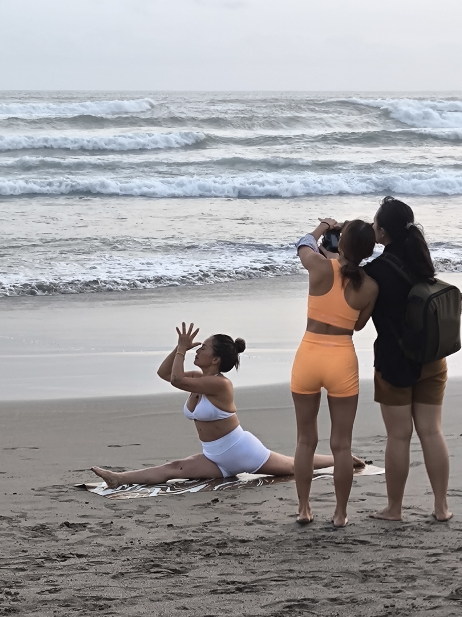 Two women stand on a beach in North Kuta, Indonesia, looking out at the ocean, while a third woman practices yoga on a mat in front of them. The yoga practitioner is positioned in a seated straddle split, with her arms raised in a prayer pose. The two women standing are facing away from the camera, and one is taking a photo with her phone. The beach has wet sand, and the ocean has moderate waves. The sky is overcast.