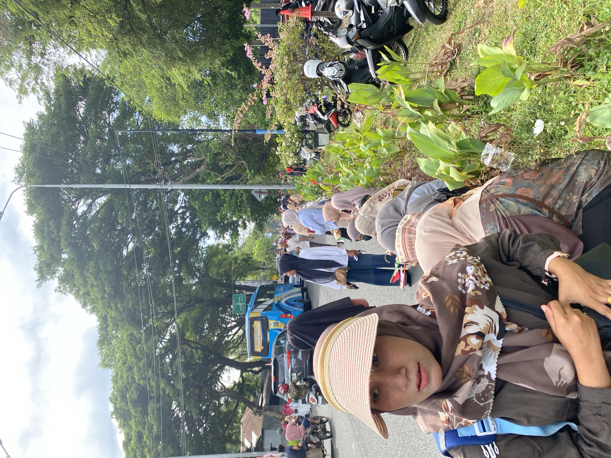 In Banda Aceh, Indonesia, on a cloudy day, a group of people are gathered by the roadside. A woman in the foreground wears a light brown hijab, a floral patterned scarf, and a straw hat. She is holding a dark bag. Behind her, other people in hijabs are seated or standing. A blue bus with the number 36 on its side is visible in the background, along with a black car and several motorcycles parked along the grassy median. Lush green trees line the street, and a street light is present on the left side of the frame. The scene appears to be a public gathering or event, possibly a procession or a religious celebration, given the attire of the individuals.