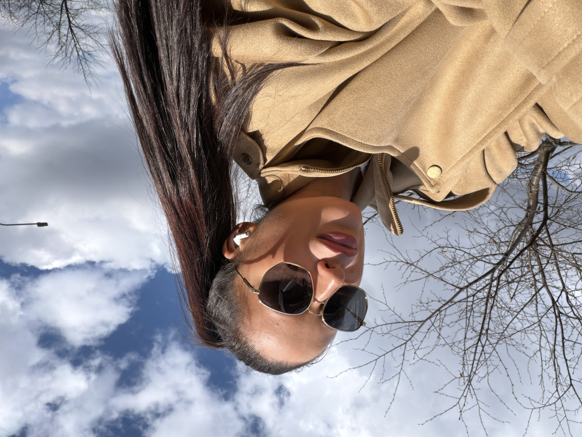 An upward-facing, portrait-style photograph, originally oriented upside down, captures a person from a low angle against a backdrop of the sky and bare trees in Khimki, Russia. The individual, likely female, has long, dark brown hair styled in a high ponytail that extends out of frame to the left. She is wearing a light brown or tan jacket, possibly made of suede or a similar textured fabric, with visible snap buttons and a zipper.
Her face is angled slightly to the right, looking upwards. She is wearing stylish hexagonal or octagonal sunglasses with dark lenses and a thin gold-toned frame. The brand "POLAROID" is visible, inverted, on the inner arm of the left lens. White wireless earbuds are visible in both ears. Her lips are slightly parted in a subtle pout.
The background primarily features a bright sky with scattered white clouds and patches of clear blue. Towards the upper right and lower left of the frame, the intricate network of bare branches from deciduous trees is visible, suggesting either late autumn, winter, or early spring. A single street light fixture can be seen in the upper left corner against the sky. The lighting is bright and natural, indicating daytime.
