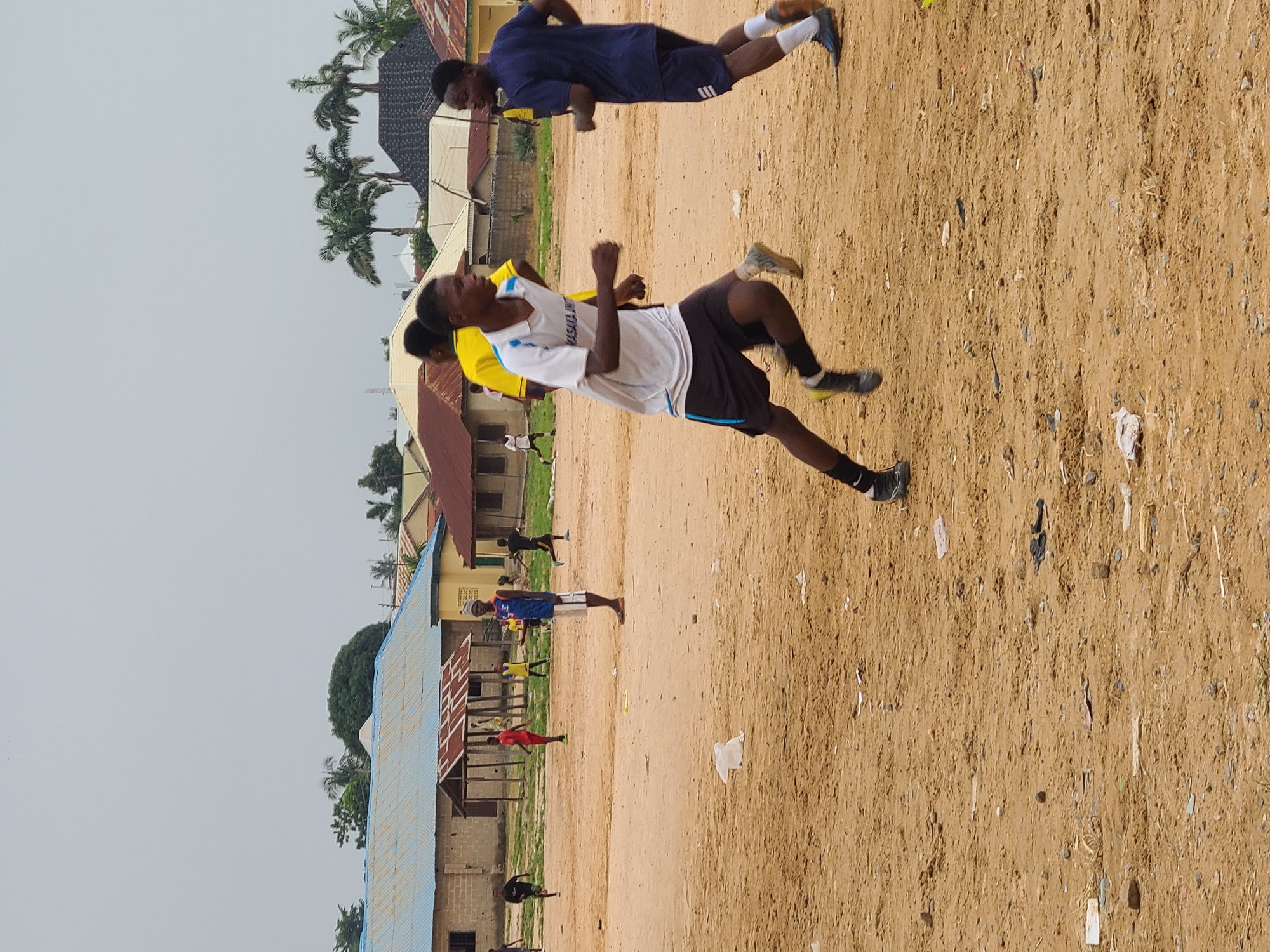 The image shows a group of young men on a dirt field, likely participating in a sports or physical training session in Masaka, Nigeria. The setting appears to be an outdoor recreational area adjacent to some buildings with corrugated iron roofs and brick structures. Palm trees are visible in the background, suggesting a tropical or subtropical climate.
The primary subjects are several young men in athletic wear. In the foreground, a young man in a white t-shirt with blue trim and black shorts is captured mid-stride, appearing to be running or performing an exercise. Another young man in a dark blue t-shirt and dark shorts is in the background, also in motion, possibly doing push-ups or a similar exercise. Other individuals in similar athletic attire can be seen in various poses on the field, suggesting an active training session.
The time of day appears to be daytime, with a bright, overcast sky dominating the upper portion of the image, indicating potentially cloudy or hazy weather. The ground is a light brown dirt, scattered with debris such as small stones, bits of paper, and plastic.
There is visible text on the white t-shirt of the foreground subject, though it is partially obscured and difficult to read completely. Some letters appear to be "MASALIKA" followed by "UNI."
The overall impression is one of organized physical activity in a rural or semi-urban Nigerian setting. The individuals' postures suggest exertion and focus on their training.