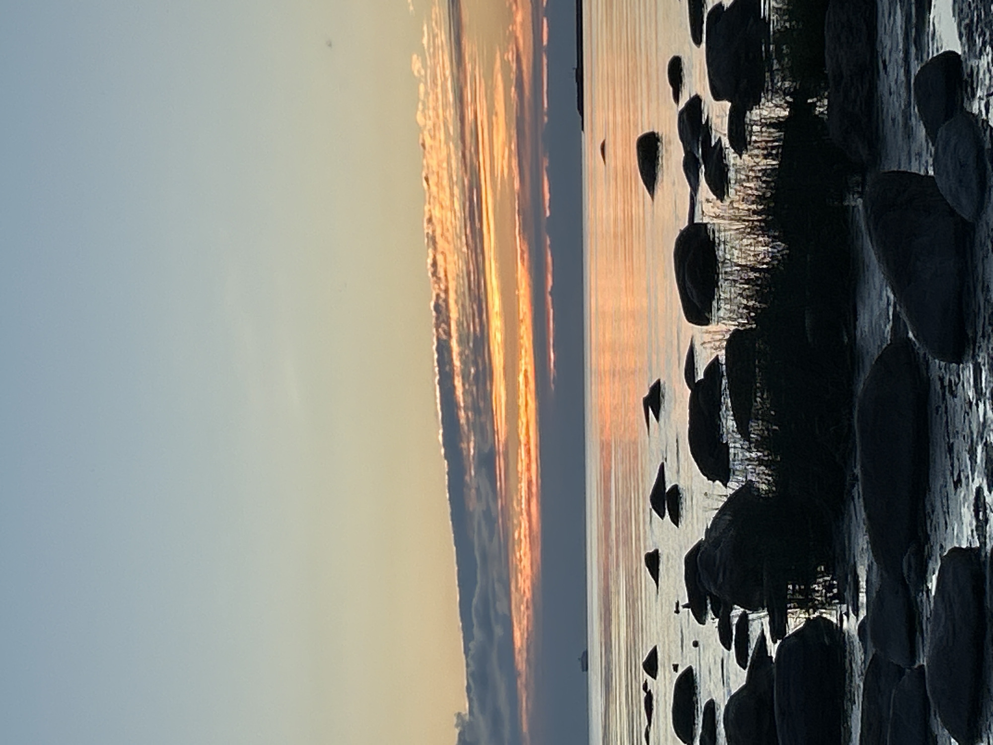 The image depicts a serene coastal scene at dusk in Haabneeme alevik, Estonia. The sky is a gradient of pale blue and soft orange, with wispy clouds catching the last rays of the setting sun. The sea is calm, reflecting the colors of the sky. Numerous dark, smooth rocks are scattered along the shoreline and in the shallow water, some partially submerged. In the distance, a faint silhouette of a ship can be seen on the horizon. The overall impression is one of tranquility and natural beauty.