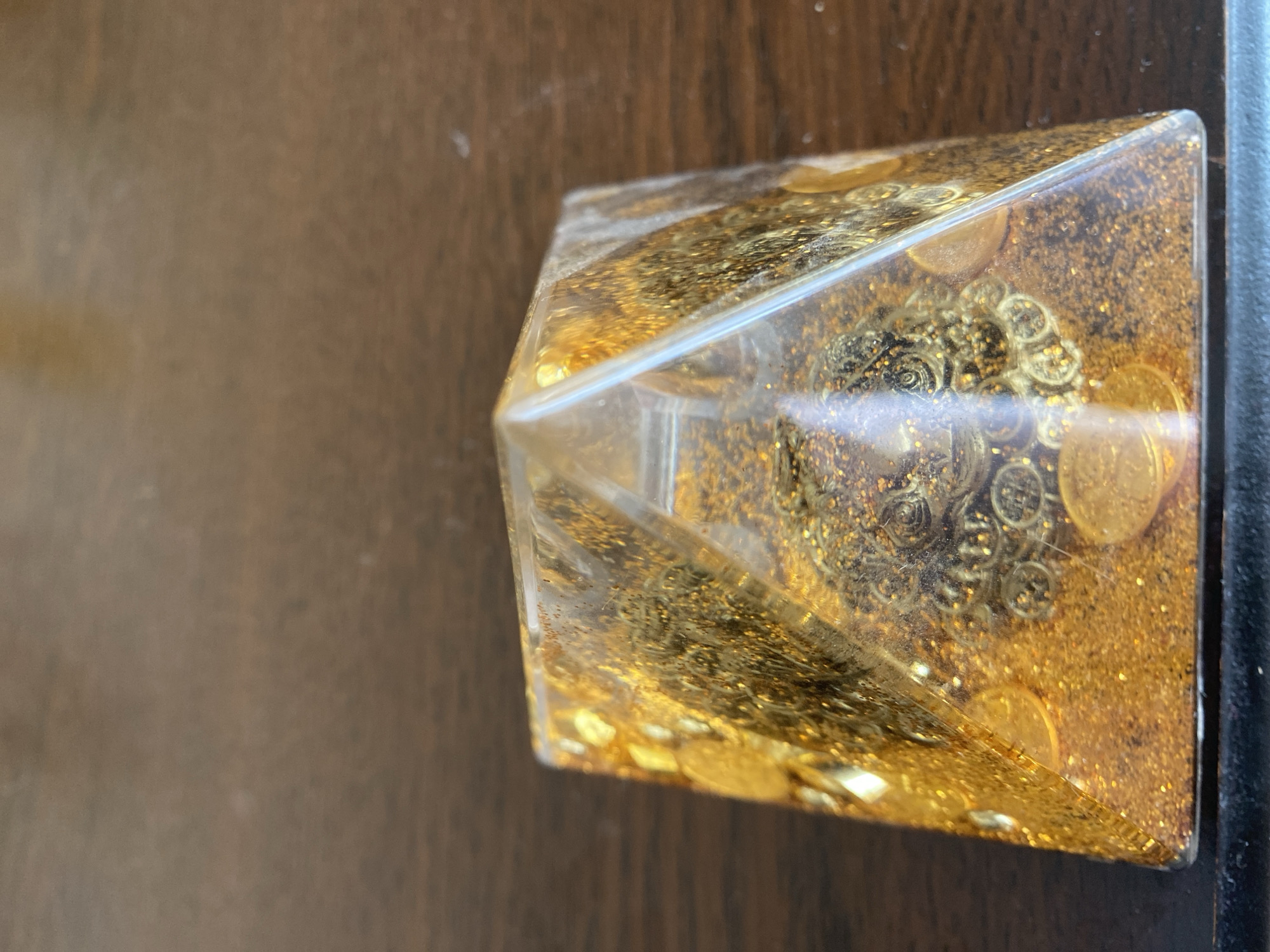 The image displays a close-up, low-angle, vertical shot of a clear pyramid-shaped object filled with gold glitter and several small, round, gold-colored coins. The pyramid is resting on a dark brown, wooden surface, which appears to be a table. The background is out of focus and consists of the same wooden surface. There are no people, animals, or any other discernible objects present. The image's lighting is warm and focused on the pyramid, highlighting the reflective qualities of the glitter and coins. The location in the city of Brest, Belarus, is not discernible from the provided image.