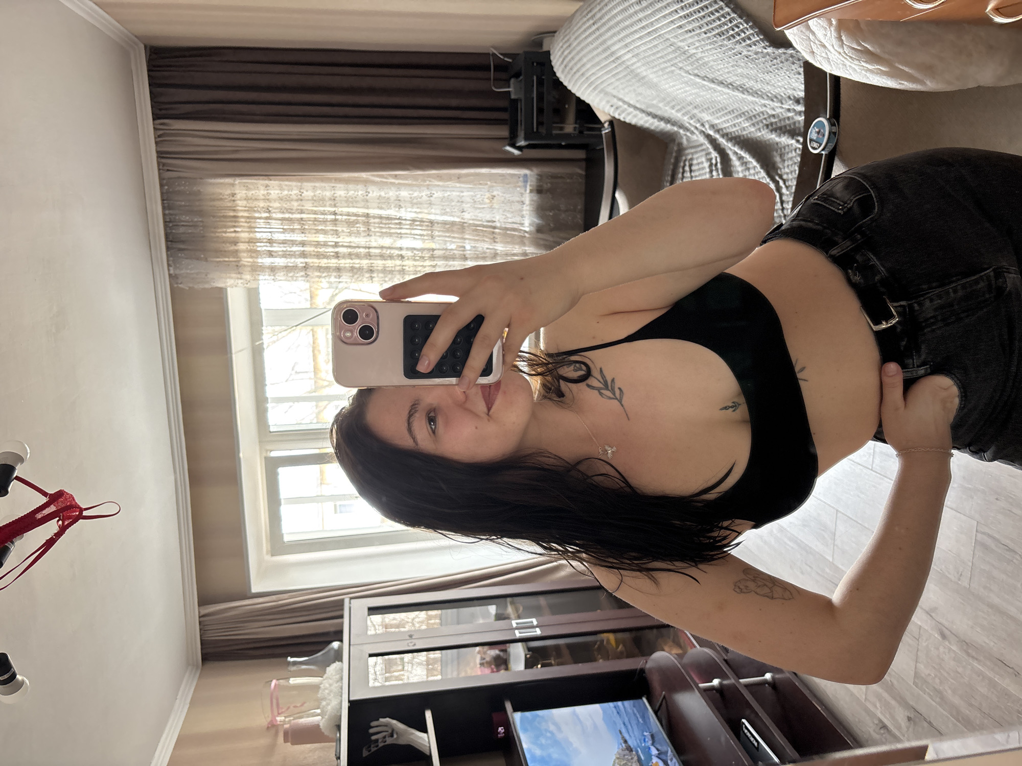 A young woman with dark hair and a fair complexion poses for a mirror selfie inside an apartment. She is wearing a black sports bra and dark jeans. A phone with a light pink case is held up, obscuring part of her face. Two tattoos are visible: one on her sternum and another on her left arm. The apartment features beige walls, brown curtains, a window, a bookcase, a chandelier, and a bed with a gray and white striped comforter. The scene is reminiscent of an apartment setting, though the location in Perm, Russia is not directly discernable from the photo.