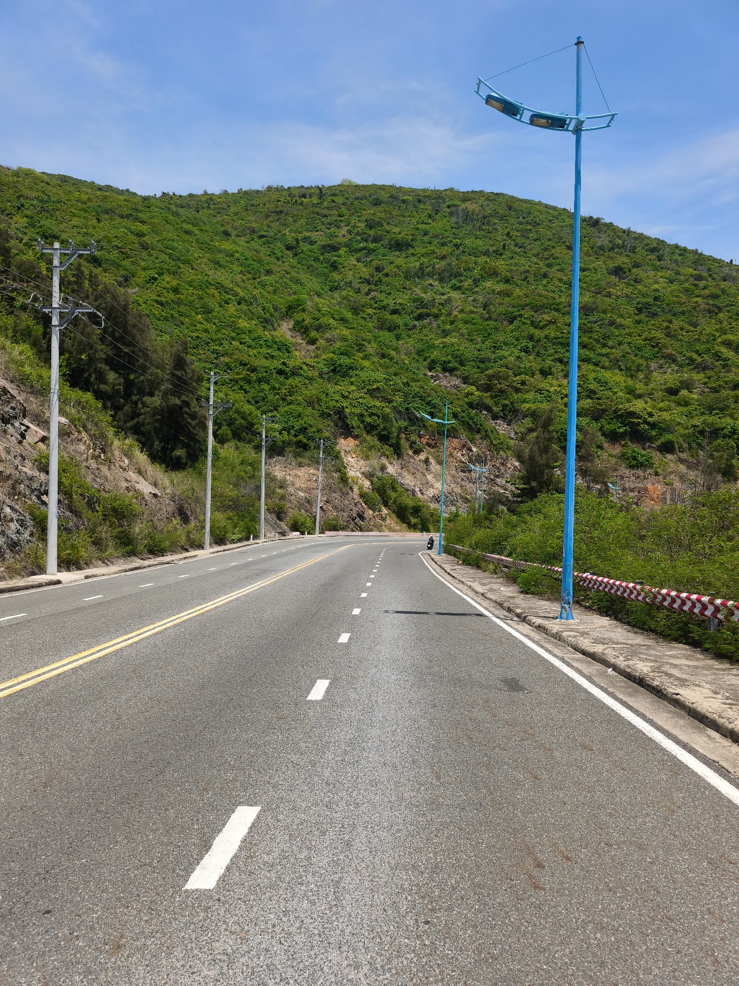 The image depicts a road winding through a mountainous area, likely in Bac Nha Trang Ward, Vietnam. The road is paved with asphalt and marked with double yellow lines and dashed white lines, indicating it is a two-lane road. On the left side of the road, a steep, rocky embankment covered with lush green vegetation rises, with utility poles and power lines running alongside it. A large blue street light stands prominently on the right side of the road, casting a shadow on the asphalt. Beyond the road, the landscape is dominated by a densely vegetated green mountain slope under a clear blue sky with scattered clouds. The right side of the road features a concrete sidewalk and a guardrail with red and white diagonal stripes. A single motorcycle is visible in the distance on the road.