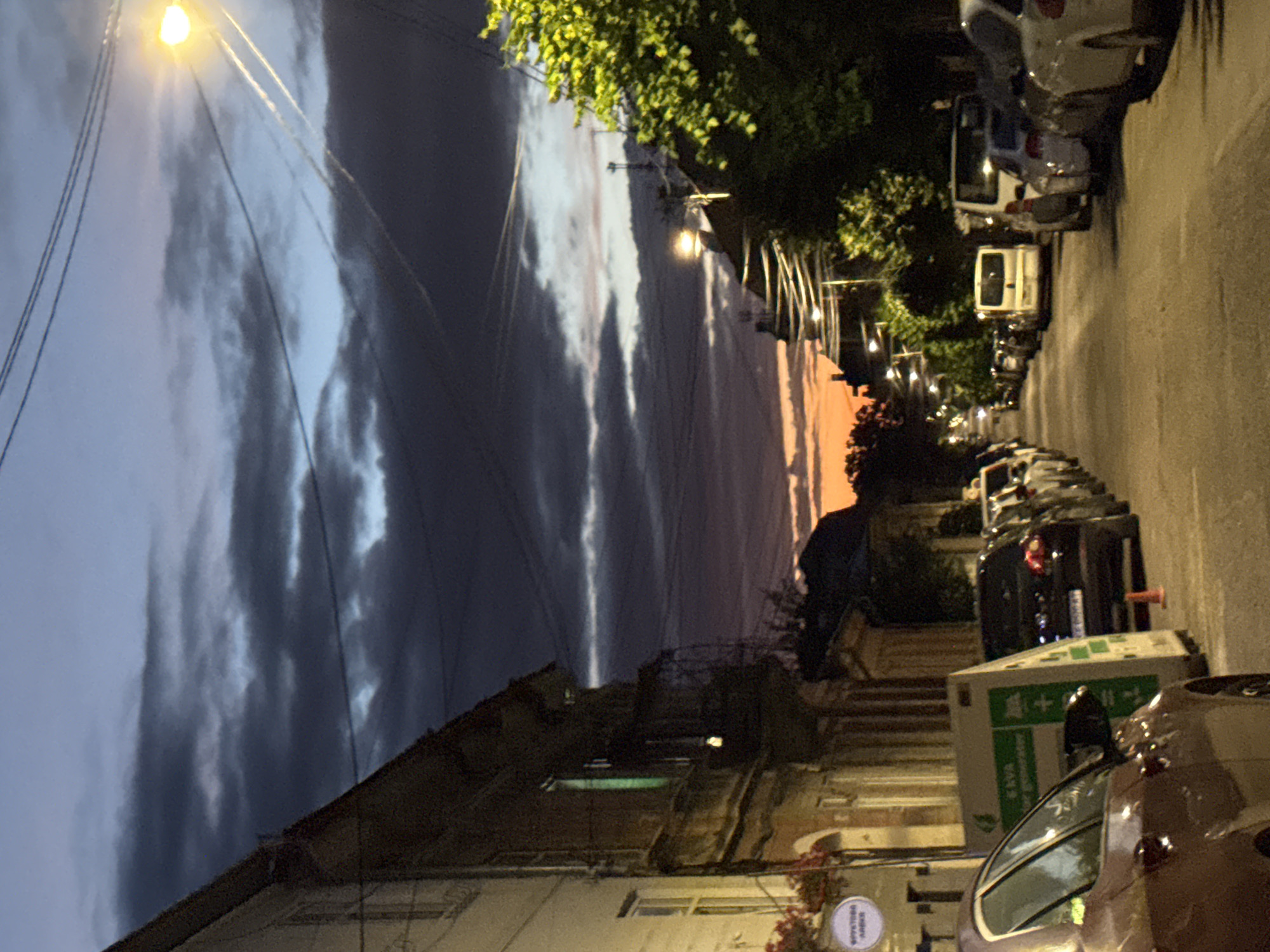 The image shows a street scene at dusk in Chernivtsi, Ukraine. The sky is filled with dramatic clouds, with a gradient of colors from deep blue to orange near the horizon. A street lamp with a bright, warm light illuminates part of the sky and street. The street is lined with parked cars, mostly sedans and SUVs, and a white van. On the left side of the street, there is a multi-story building with traditional architecture, including balconies and arched windows. A small sign with Cyrillic text is visible on the building. On the right side of the street, trees with lush green foliage are present, partially obscuring other buildings and streetlights. The overall atmosphere is one of a quiet evening in a European city.