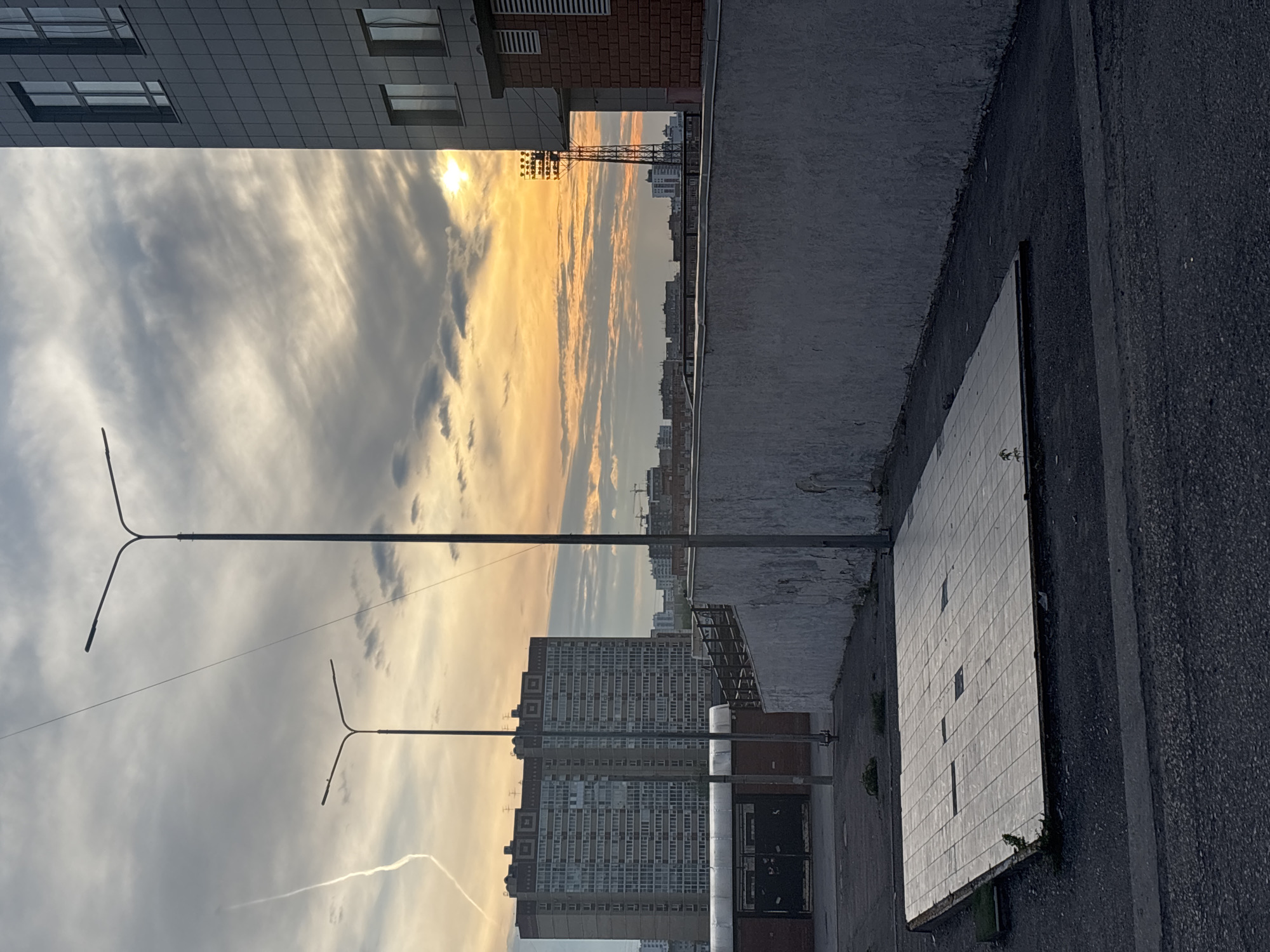This image captures a cityscape, likely in Perm, Russia. The view is oriented to the side, showing buildings silhouetted against a sunset sky. Streetlights stand in the foreground against the vibrant sky. A gray concrete wall is in the bottom right, partially covered in what appears to be white tiles. Behind the wall and extending into the distance is a row of buildings. The sky is filled with a mixture of gray and golden clouds, indicating the presence of sunlight.