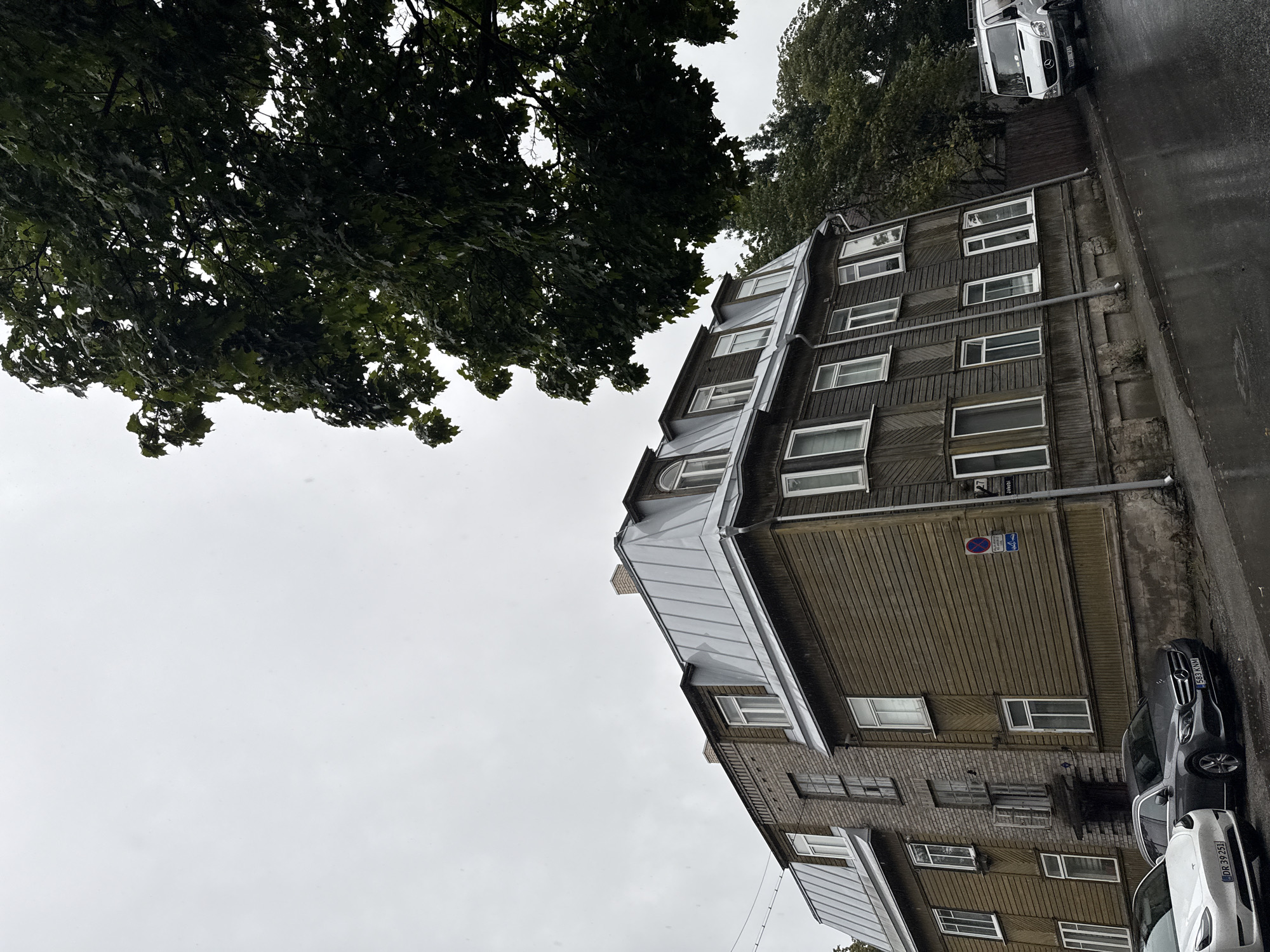The image shows a street scene in Tallinn, Estonia, on a cloudy, rainy day. A tall, old wooden building with a metal roof dominates the left and center of the frame. The building has multiple floors with numerous windows, some of which are dormer windows on the mansard roof. A brick section is visible on the left side of the building, attached to the wooden structure. Two cars are parked in front of the building on the wet, asphalt street. The car on the left is a white Tesla Model 3, and the car on the right is a dark gray Mercedes-Benz sedan. Further down the street, on the right side, a white Mercedes-Benz van is parked. A large tree with dark green leaves extends into the upper right portion of the image, its branches partially obscuring the sky. The street appears to be sloped. Several signs are visible on the building, including a blue and white circular sign with a red border and a number 27 on a pole.
