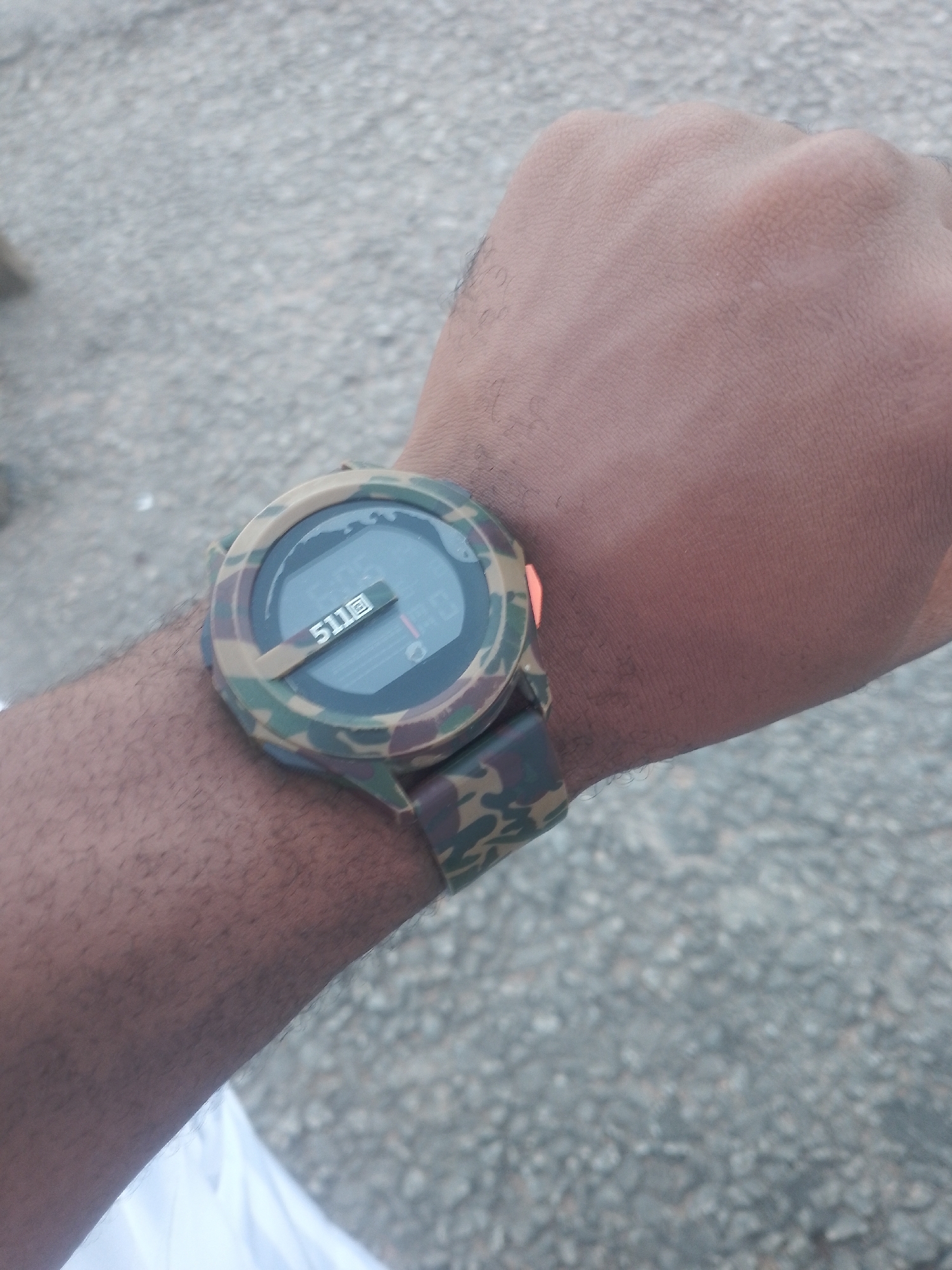 A person's wrist and forearm are visible in the foreground, with a camouflage-patterned digital watch on their wrist. The watch face displays "511B" and what appears to be time information. The background consists of a grey, gravelly surface, suggesting an outdoor or urban environment. The lighting is moderate, casting no strong shadows.
