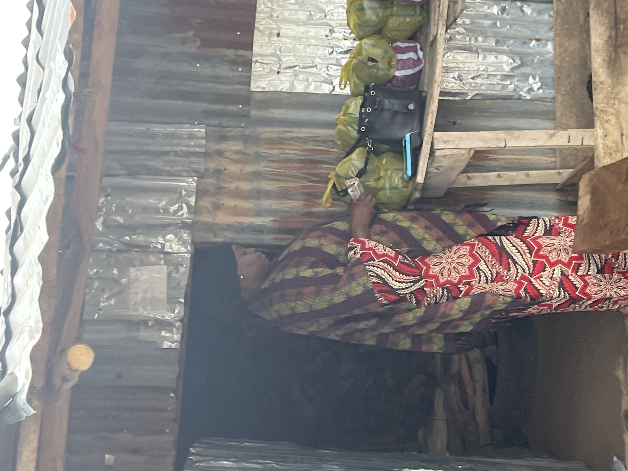 A woman stands at a stall constructed of corrugated metal in what appears to be a market in Jos, Nigeria. She is wearing a dark headscarf, a patterned shawl in shades of maroon, green, and brown, and a vibrant red and white patterned dress. Her right hand is holding some currency. On the wooden counter in front of her are several yellow plastic bags, some containing produce, and a black handbag. A small blue electronic device, possibly a point-of-sale terminal, is also on the counter. The stall is open to the elements, with a corrugated metal roof supported by wooden beams. The background appears to be the dark interior of a more enclosed space within the stall. The ground is dirt.