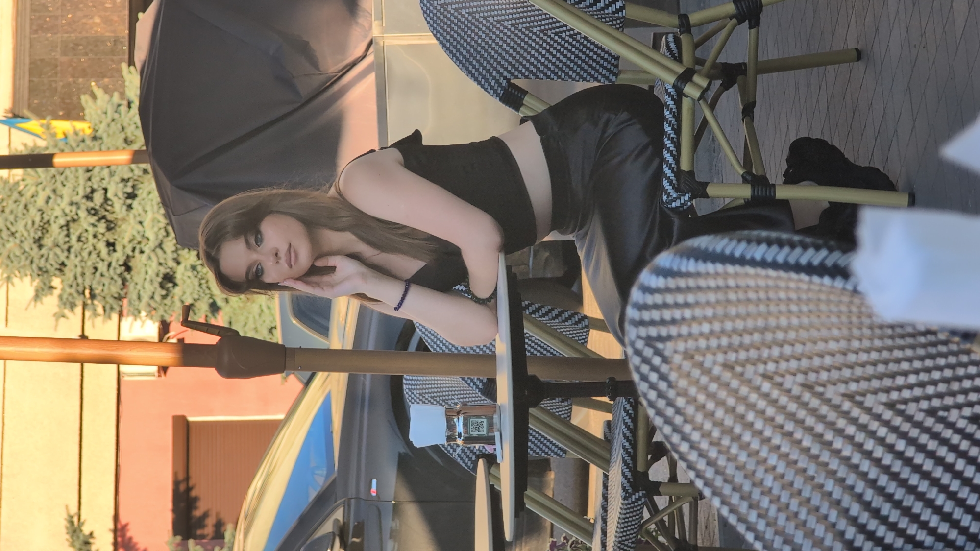 A young woman is captured in a photograph, posing outdoors at a table. She is leaning forward, resting her head on her hand, with her gaze directed towards the camera. She is wearing a black crop top and black pants, with a beaded bracelet on her left wrist. The scene appears to be an outdoor seating area, possibly a cafe or restaurant, with chairs and tables set up. A large black umbrella is visible in the background, suggesting it might be sunny and the umbrella is for shade. The overall mood seems relaxed and stylish, possibly a casual photoshoot or a moment captured during an outing. The lighting suggests it could be daytime, likely afternoon, due to the warm tones and shadows.