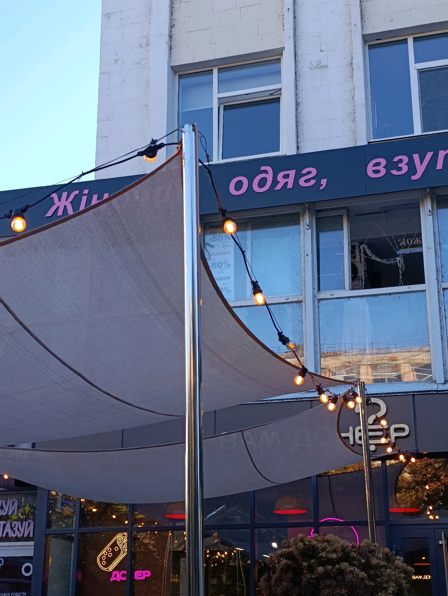 The image shows a storefront with a canopy and string lights. The storefront has large windows and a sign that says "Жіночий одяг, взуття" (Women's clothing, shoes) in pink letters. Below the main sign, there are smaller signs advertising discounts of 50%. On the left side of the image, there is a neon sign that appears to say "ДЕСЕРТ" (Dessert) in yellow and pink. The building is made of white brick. The photograph was taken outdoors during daylight, possibly late afternoon or early evening, as indicated by the warm glow of the string lights. The environment appears to be an urban setting, possibly a commercial street in Cherkasy, Ukraine, given the Cyrillic lettering.