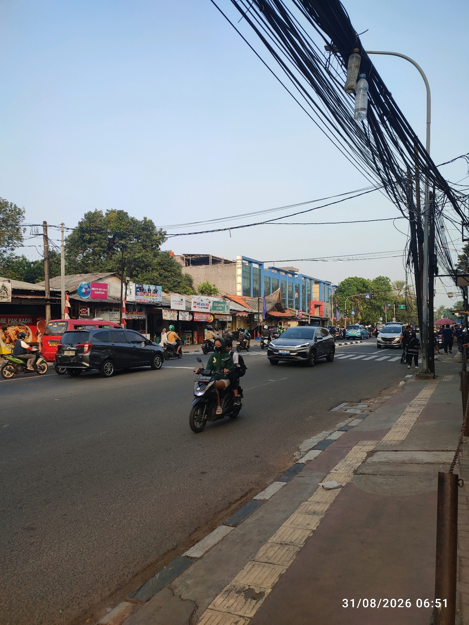 The image depicts a street scene in East Jakarta, Indonesia, on August 31, 2026, at 06:51. The foreground shows a two-lane asphalt road with moderate traffic. A dark-colored SUV is in the left lane, heading away from the viewer, and a scooter with two occupants is in the right lane, also moving away. On the right side of the road, a concrete sidewalk is visible, with a metal pole and a chain. Buildings line the left side of the street, including storefronts with various signs, some in Indonesian. Trees are present, and a complex network of overhead electrical wires crisscrosses the sky. The sky is clear and blue, indicating it is daytime, likely early morning due to the timestamp.