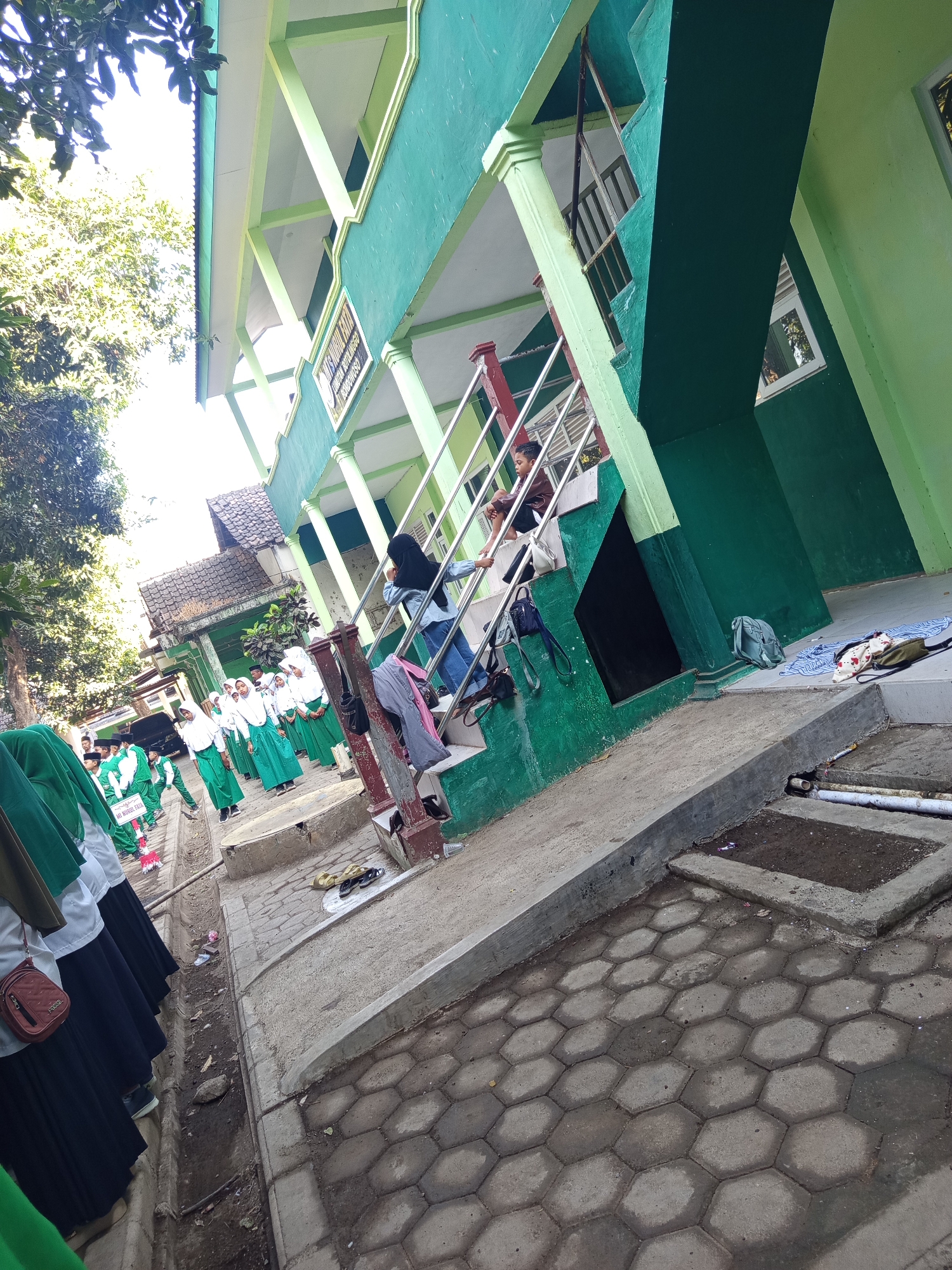 The image depicts an outdoor scene at what appears to be a school building in Lumutan, Indonesia. The building has a distinct green facade with white and light green accents. A staircase with a metal railing, painted red and silver, leads up to the upper levels of the building. Several students, predominantly wearing green hijabs and white or green uniforms, are gathered near the base of the staircase and along a walkway. Some students are ascending the stairs, while others are standing and conversing. One student is seated on the staircase. Various objects, including bags, a blanket, and footwear, are scattered around the area. Trees and foliage are visible in the background, suggesting a bright, sunny day. The ground is paved with hexagonal concrete blocks and features a drainage ditch. A sign with Indonesian text is partially visible. The overall impression is of a typical school day scene in a tropical environment.