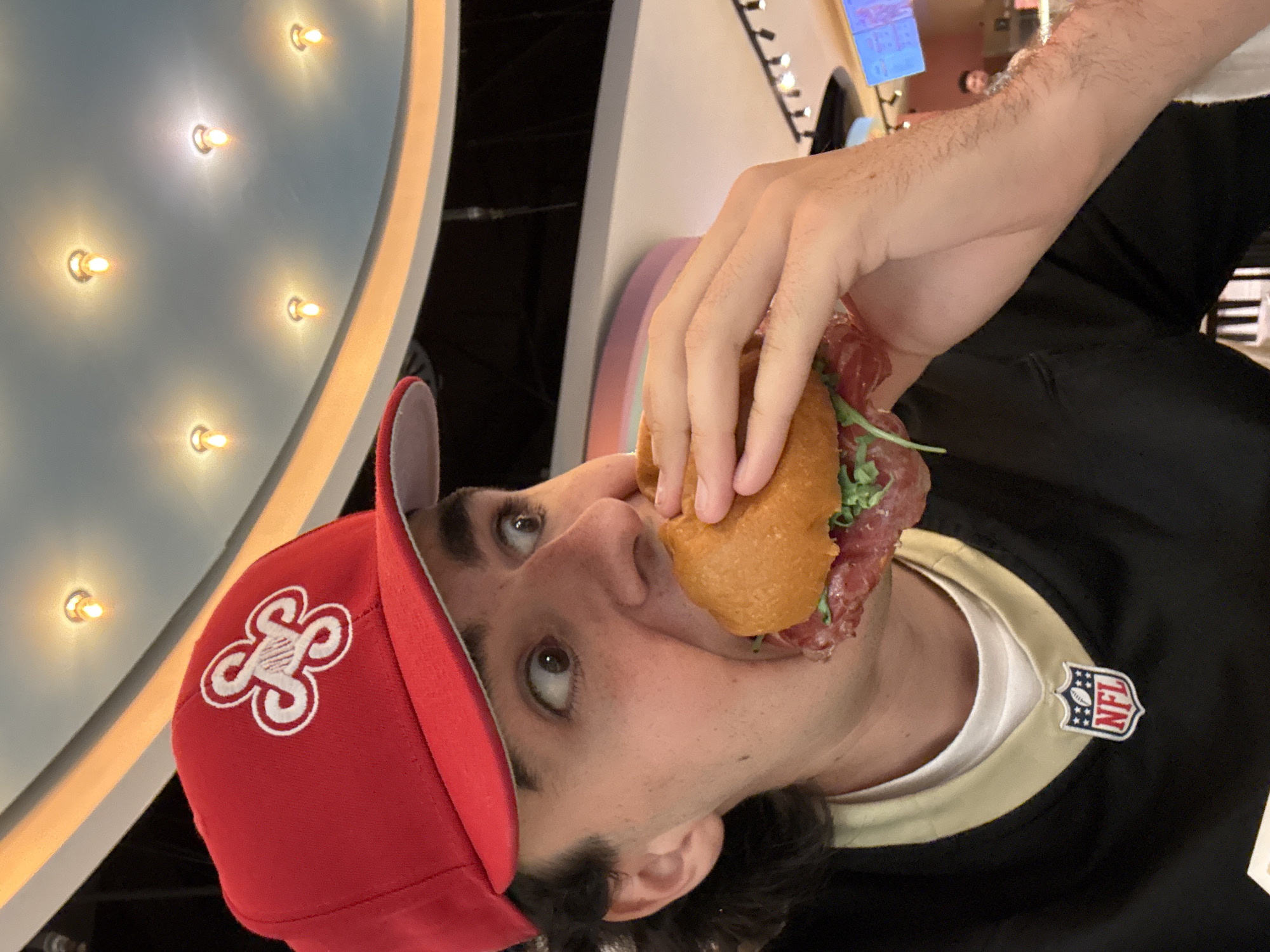 A person is eating a sandwich indoors, appearing to be in a restaurant or food establishment. The person is wearing a red baseball cap with a white clover-like logo and a black and gold shirt with an NFL logo. The background includes a wall with embedded lights and other patrons in the distance. Based on the hat and the shirt, it seems that the person is a New Orleans Saints fan. The city of Ferno, Italy cannot be determined from this image.