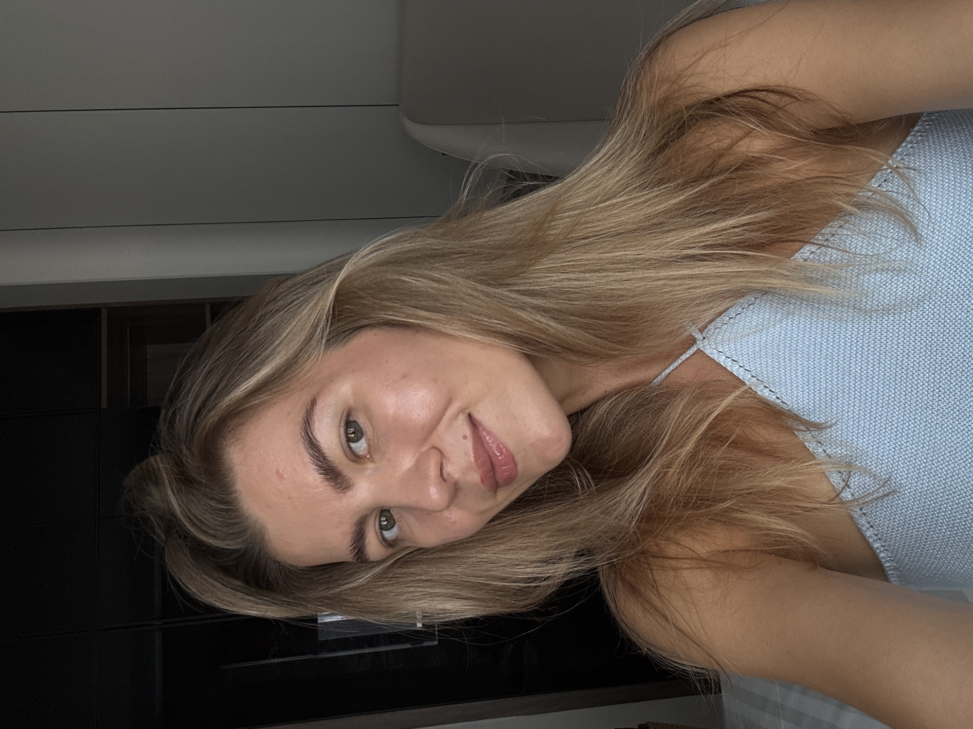 A woman with blonde hair and fair skin is facing the camera and smiling slightly. She is wearing a light blue top. The background appears to be an interior space, possibly a kitchen, with a white wall or cabinet above and black cabinets below. The location is Unknown, Unknown.