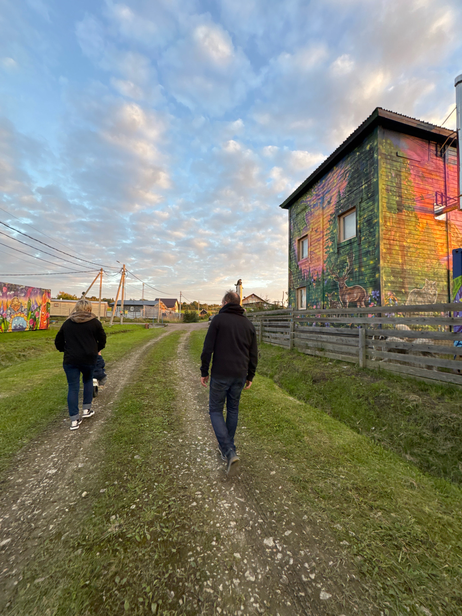 Two individuals are walking away from the camera on a dirt path in a rural area. The person in the foreground on the right is wearing a dark hooded jacket and blue jeans. The person in the foreground on the left is wearing a dark hooded jacket and blue jeans, with a child on their back, also wearing a dark jacket. They are walking towards a colorful building on the right side of the image, which is covered in murals depicting nature scenes and animals. A wooden fence runs along the side of the building, with what appear to be sheep behind it. In the background, there are several houses and utility poles with electrical wires, suggesting a small village or settlement. The sky is partly cloudy with soft light, indicating it might be early morning or late afternoon. The location is described as the city of Kempelevo, Russia.